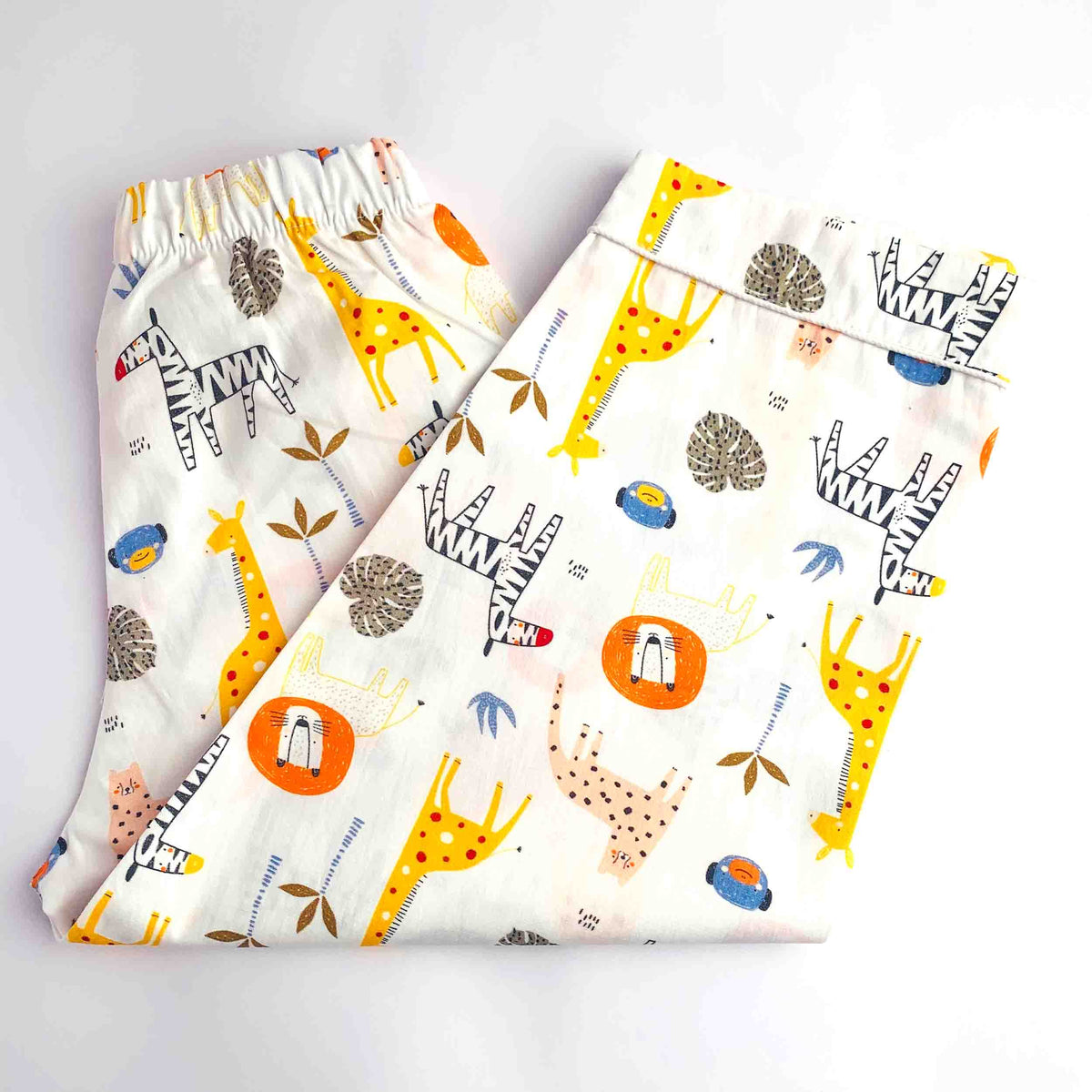 A pair of folded white children's pants with an elastic waistband, featuring a playful safari-themed print of giraffes, zebras, acacia trees, and suns in yellow, black, orange, and brown.