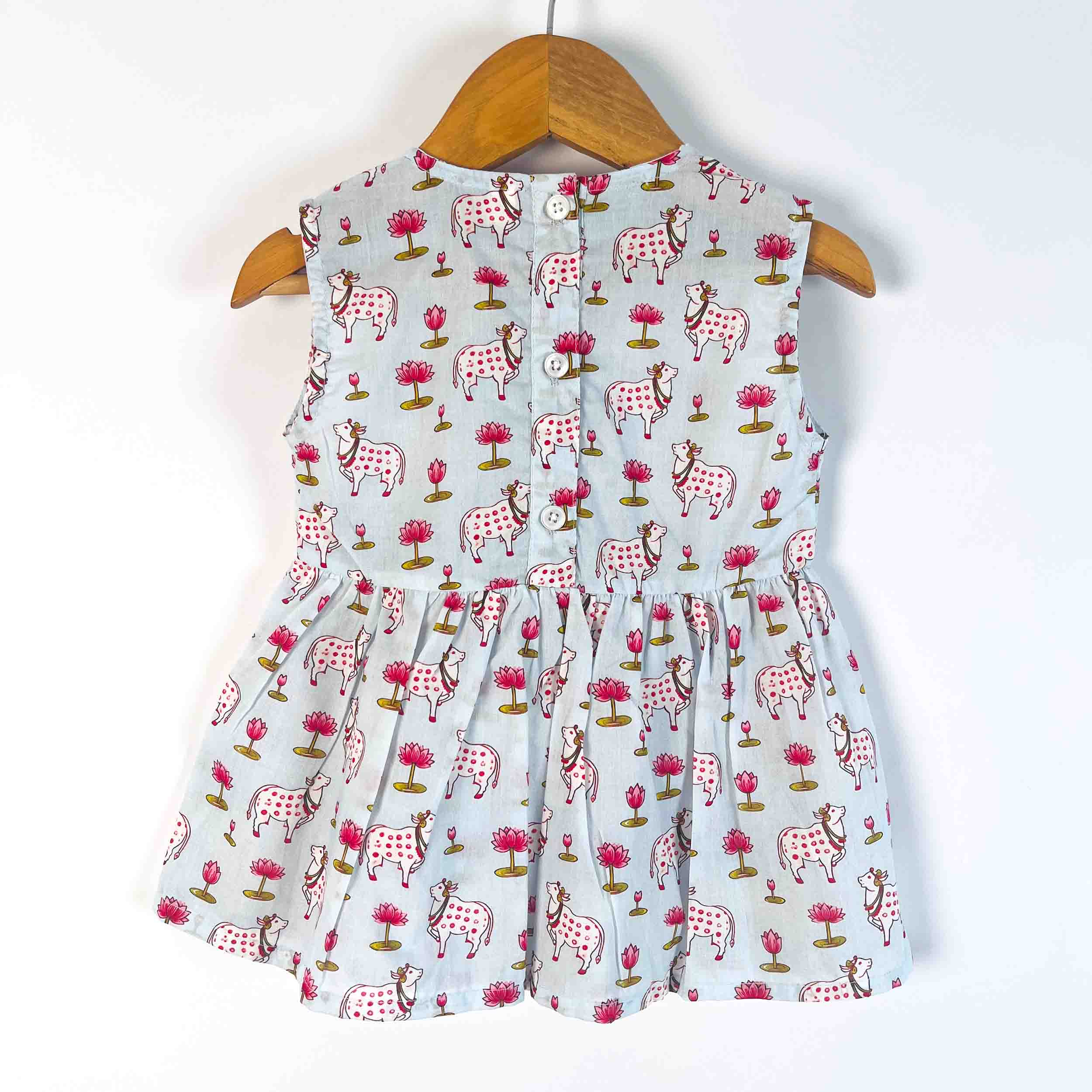 A sleeveless baby dress with a light blue-gray background and an all-over print of dalmatian dogs and pink flowers, hanging on a wooden hanger against a white wall.