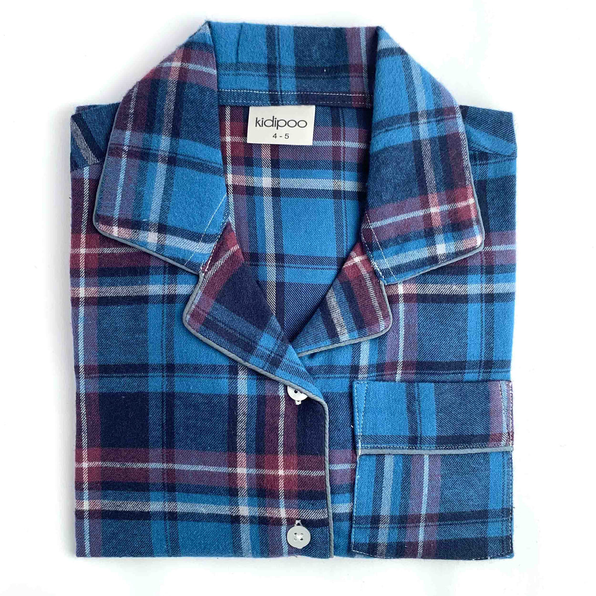 A folded children's long-sleeve pajama shirt with a blue, red, and navy plaid pattern, featuring a collar, button-down front, and a pocket on the left chest. The brand label 'kidipoo' is visible inside the collar.