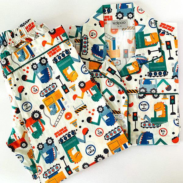 A set of children's pajamas with a dinosaur theme, including a long sleeve, front button shirt and coordinated pajamas with elastic at the waist, laid out on a white surface.