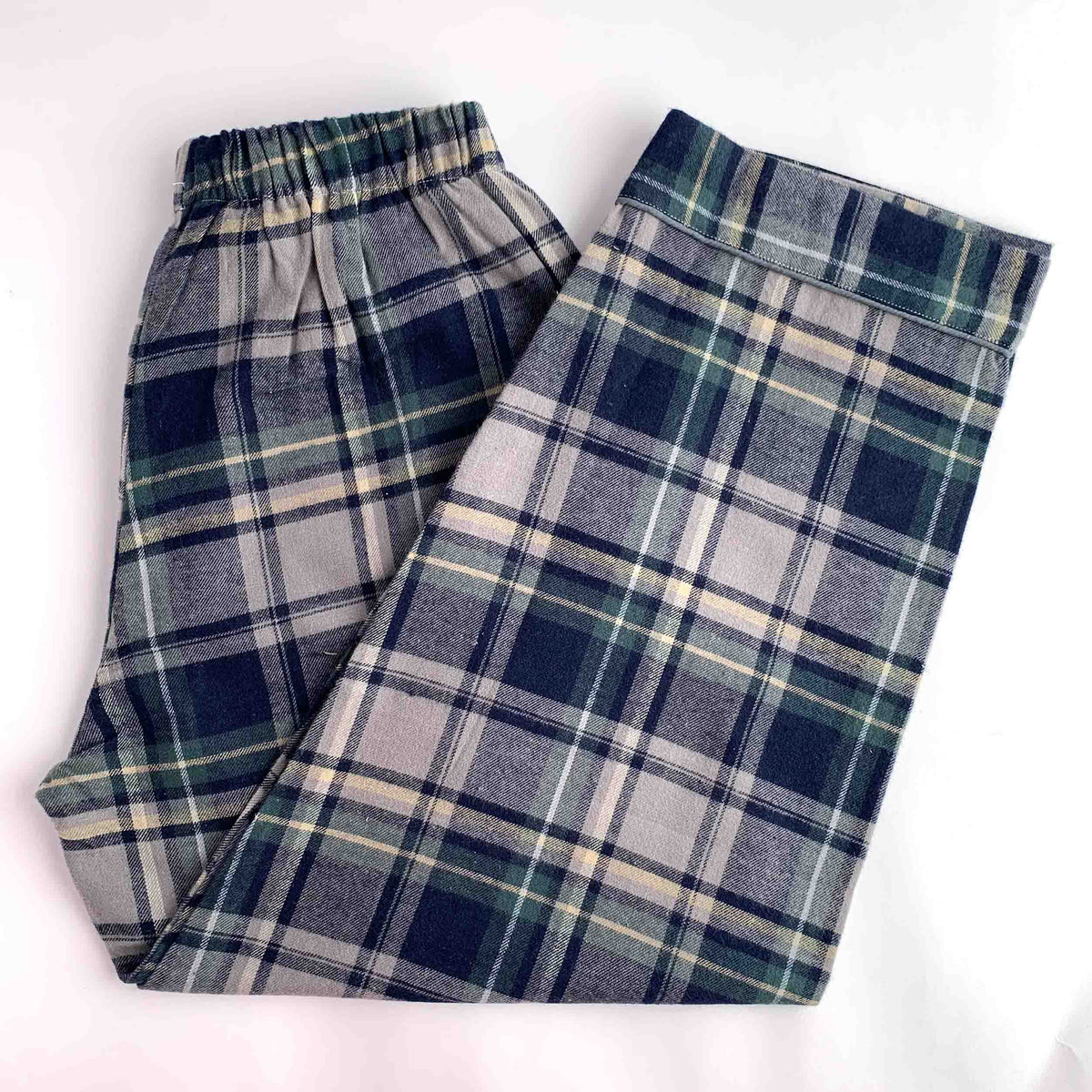 A pair of plaid lounge pants laid flat on a white background. The pants feature an elastic waistband and a classic tartan pattern in navy blue, gray, white, and thin green stripes.