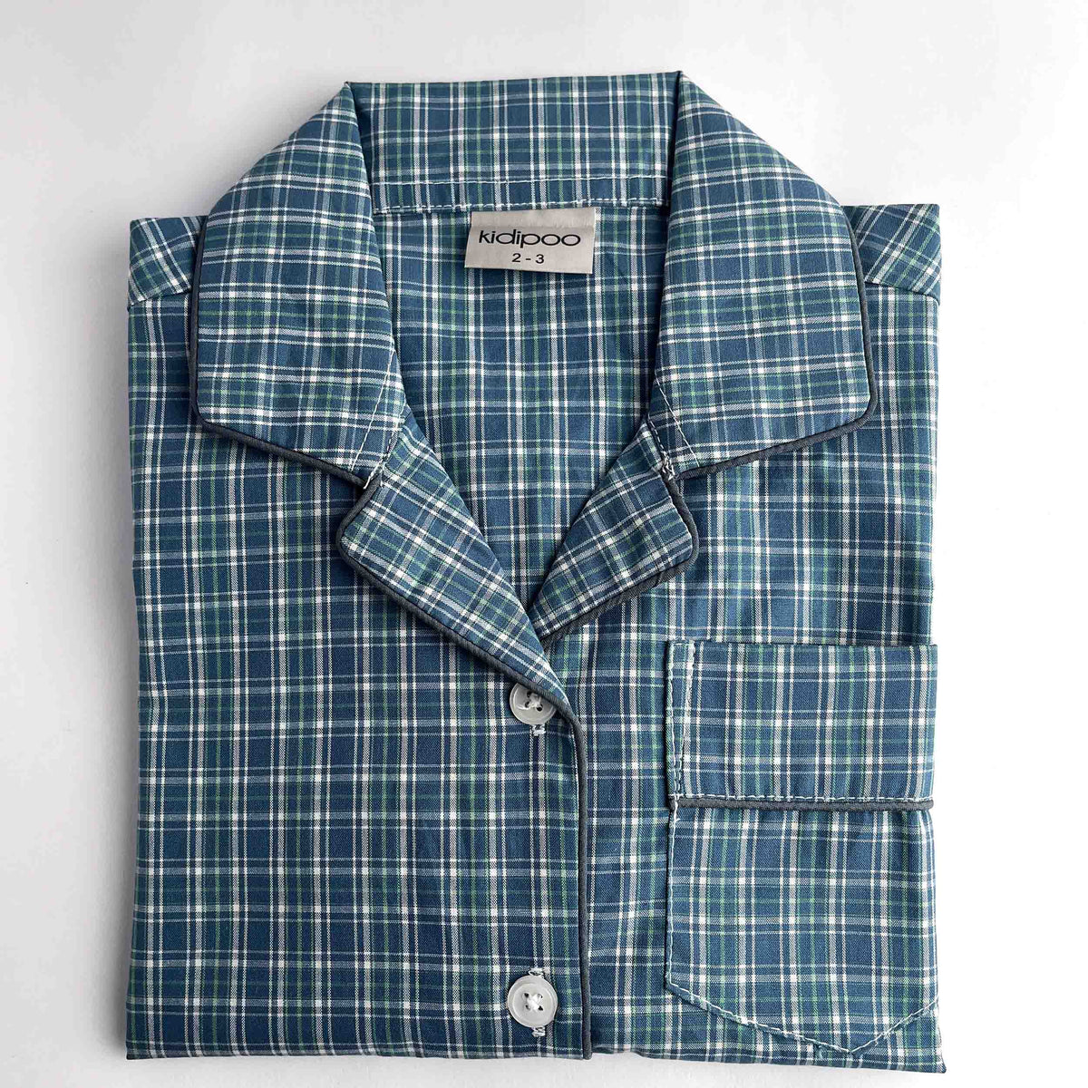 A folded long-sleeve pajama shirt with a blue, green, and white plaid pattern, featuring a collar, button front, and a chest pocket. The brand label 'kidpoo' is visible inside the collar.