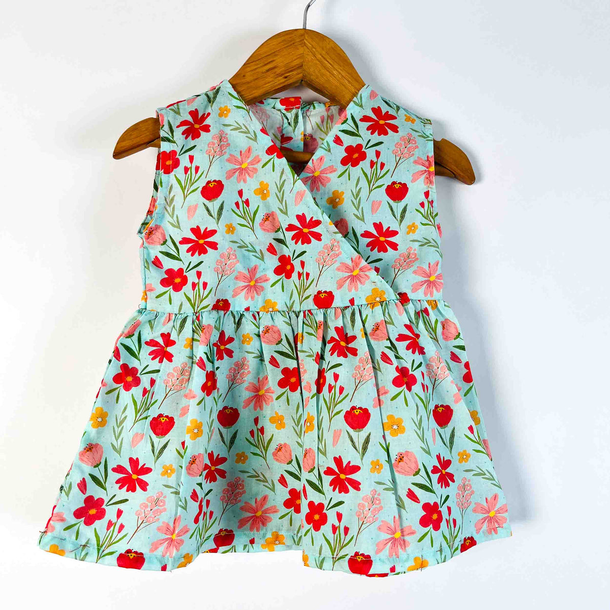 A sleeveless wrap-style dress for toddlers or girls, featuring a vibrant floral print on a light mint green background. The dress is displayed on a wooden hanger against a plain white wall.