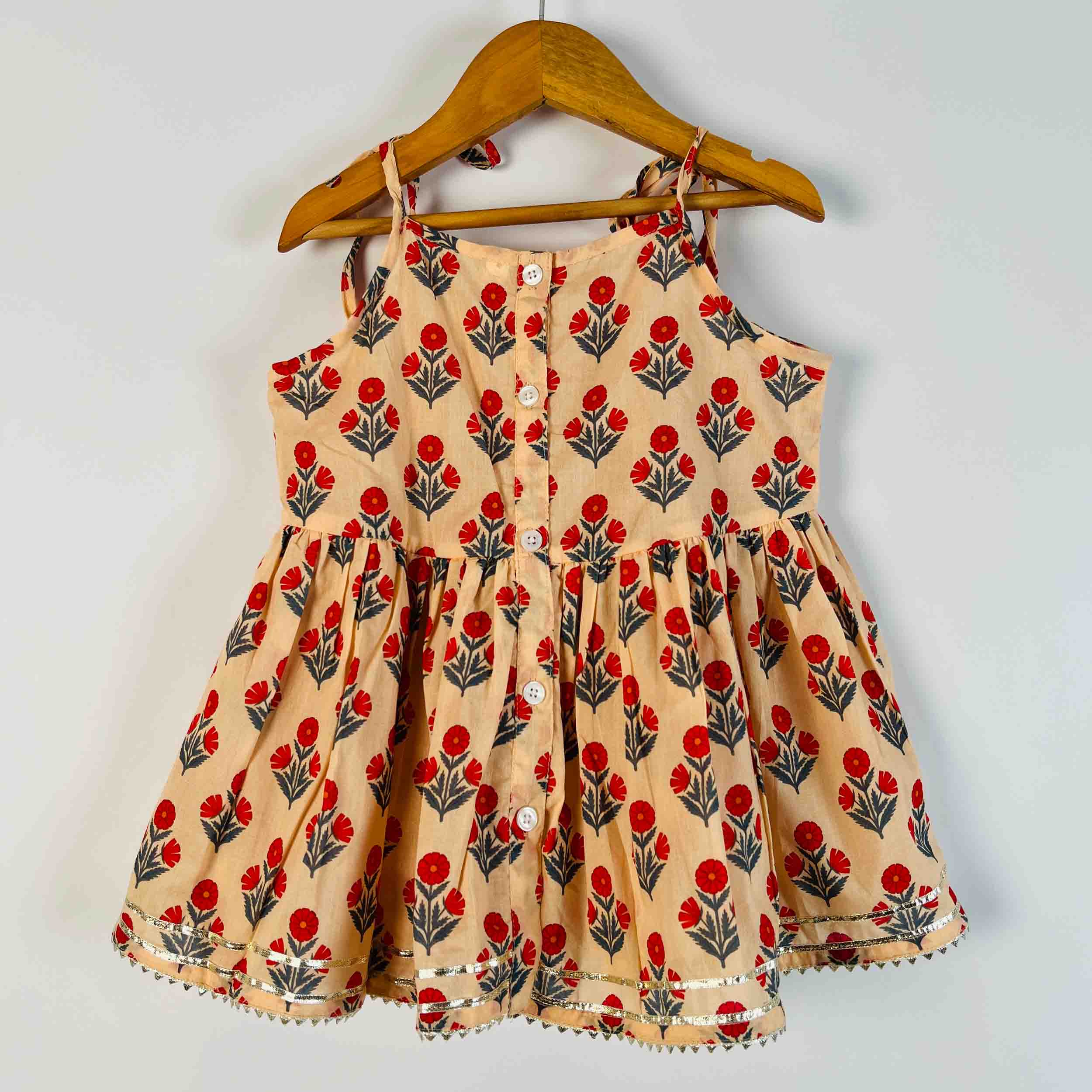 A sleeveless children's dress with a red and green floral block print on a beige background, hanging on a wooden hanger against a plain light gray wall.