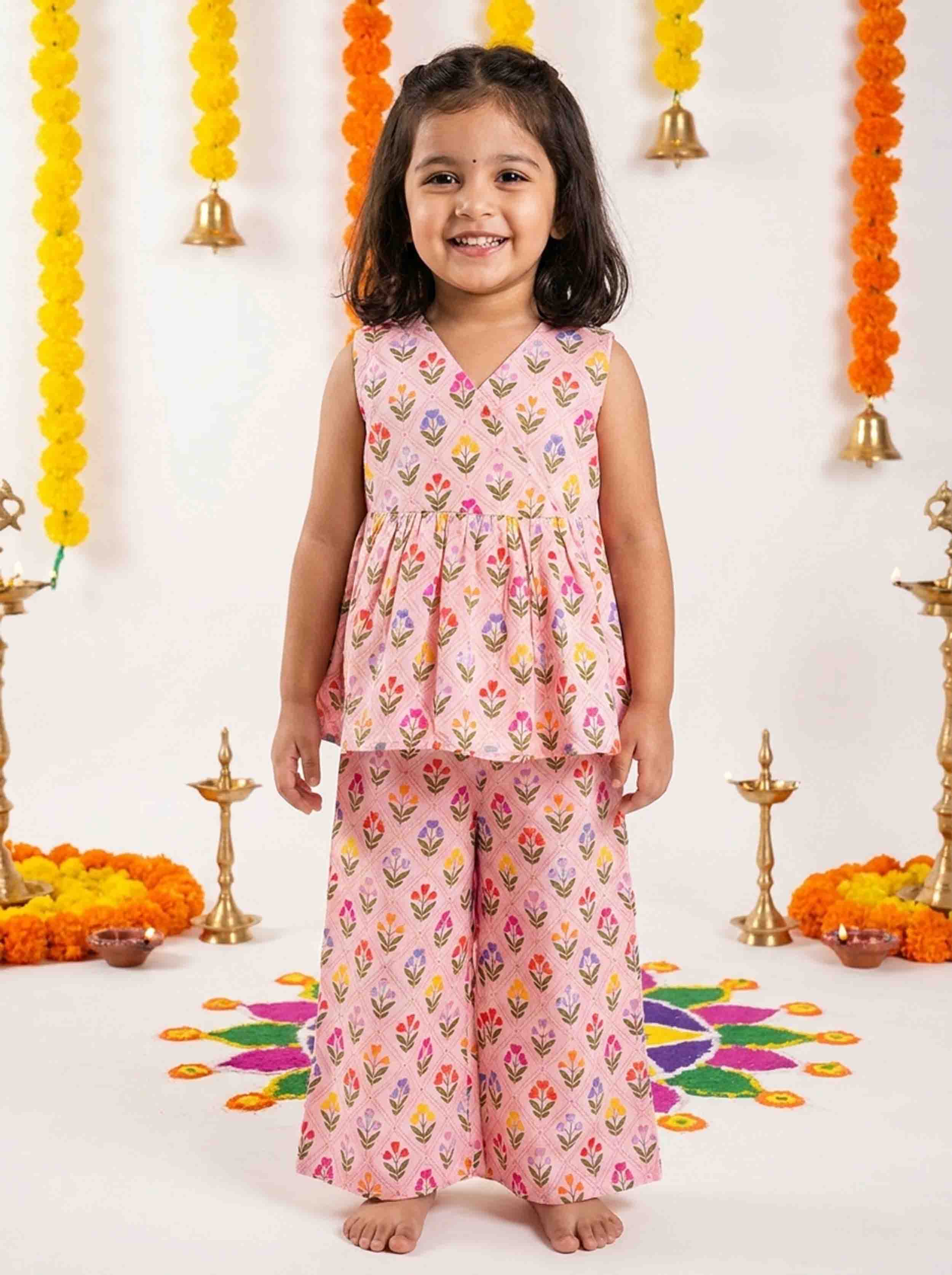 A young girl stands barefoot wearing a pink sleeveless kurta set with a colorful floral print, posed in front of a decorated white wall with marigold garlands and brass lamps.