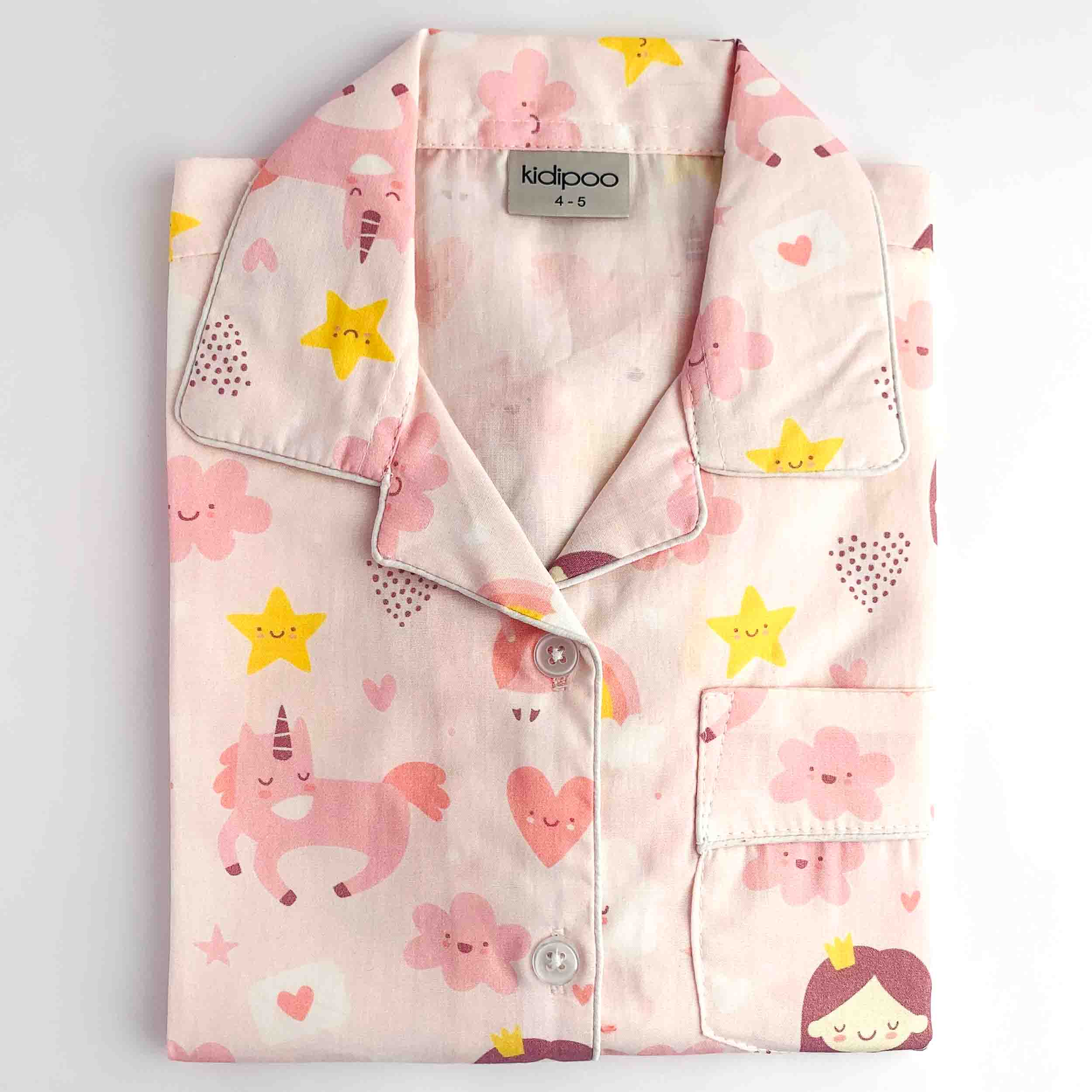 A folded children's pajama shirt with a pink base and a playful cartoon print featuring unicorns, smiling stars, hearts, and flowers. The shirt has a collar, button front, and a chest pocket.