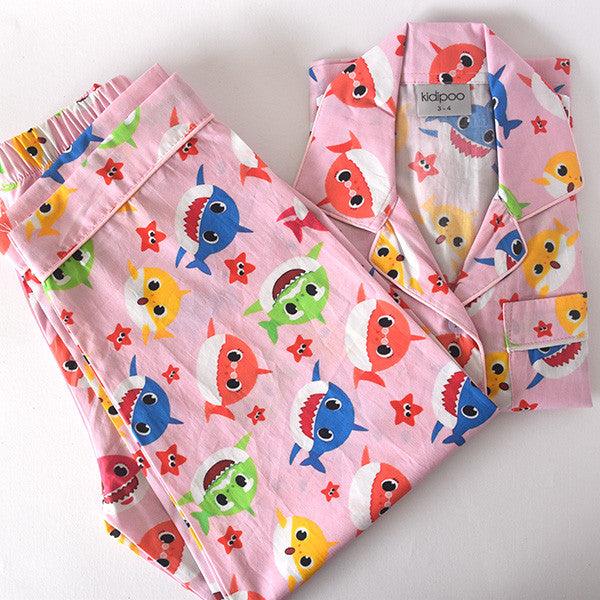 A children's pajama set with long sleeves, featuring a pink background with colorful shark characters and star patterns.
