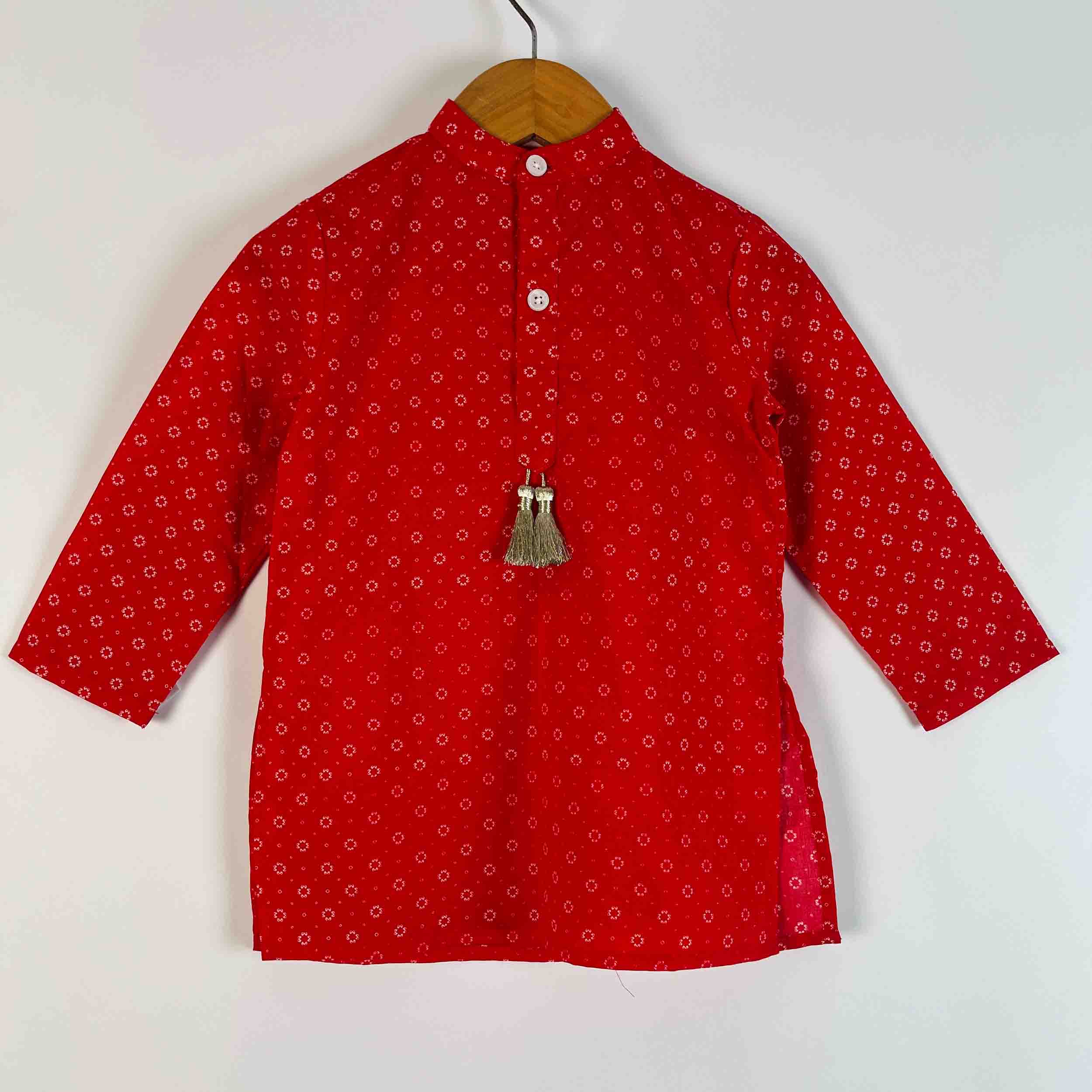A red long-sleeve kurta with a small white geometric pattern, hanging on a wooden hanger against a plain white background. It features a mandarin collar, three white buttons, and a decorative silver tassel.