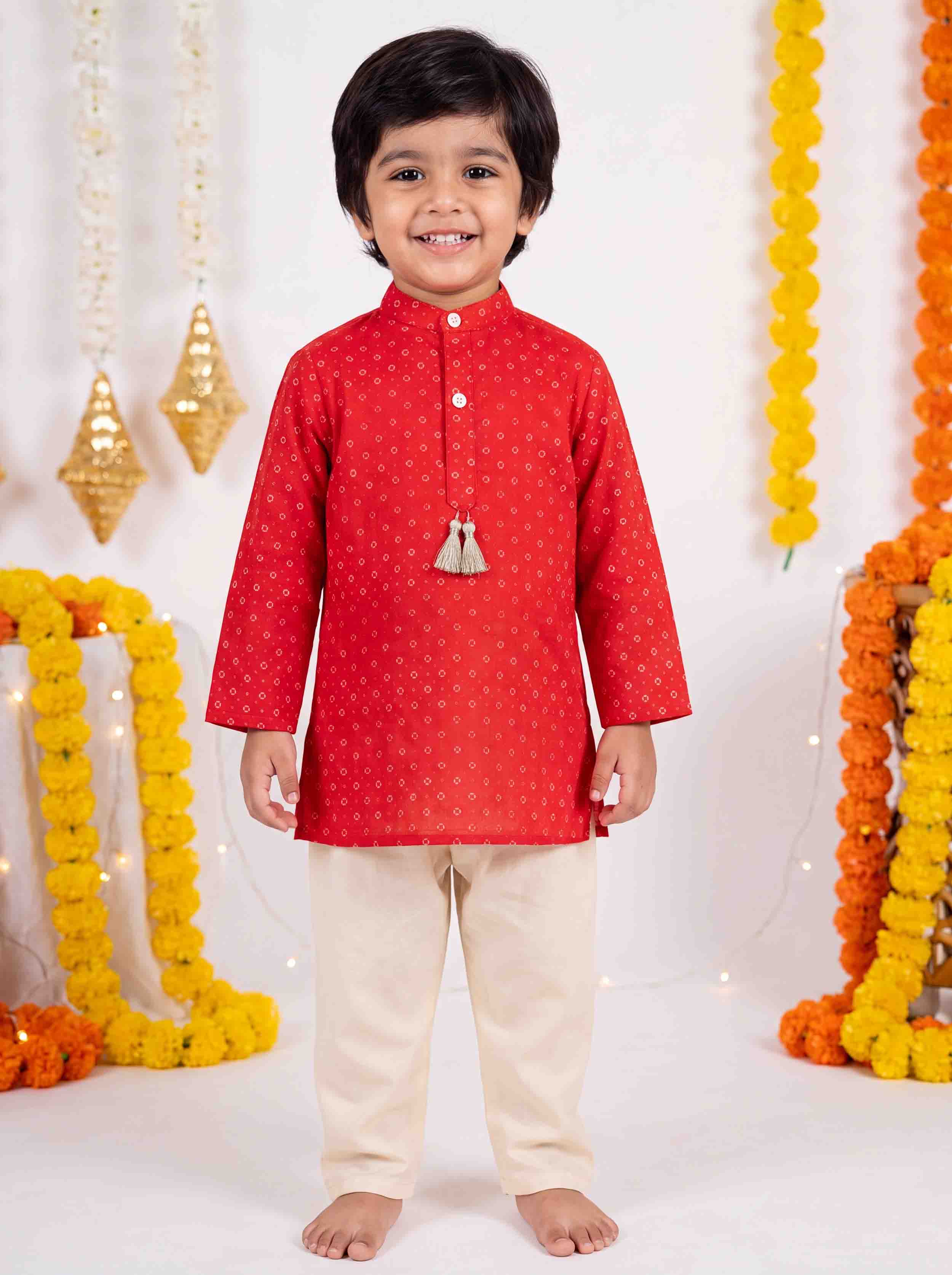 A young boy wearing a red kurta with small gold motifs and cream trousers, standing barefoot in front of a decorated white backdrop with marigold garlands.