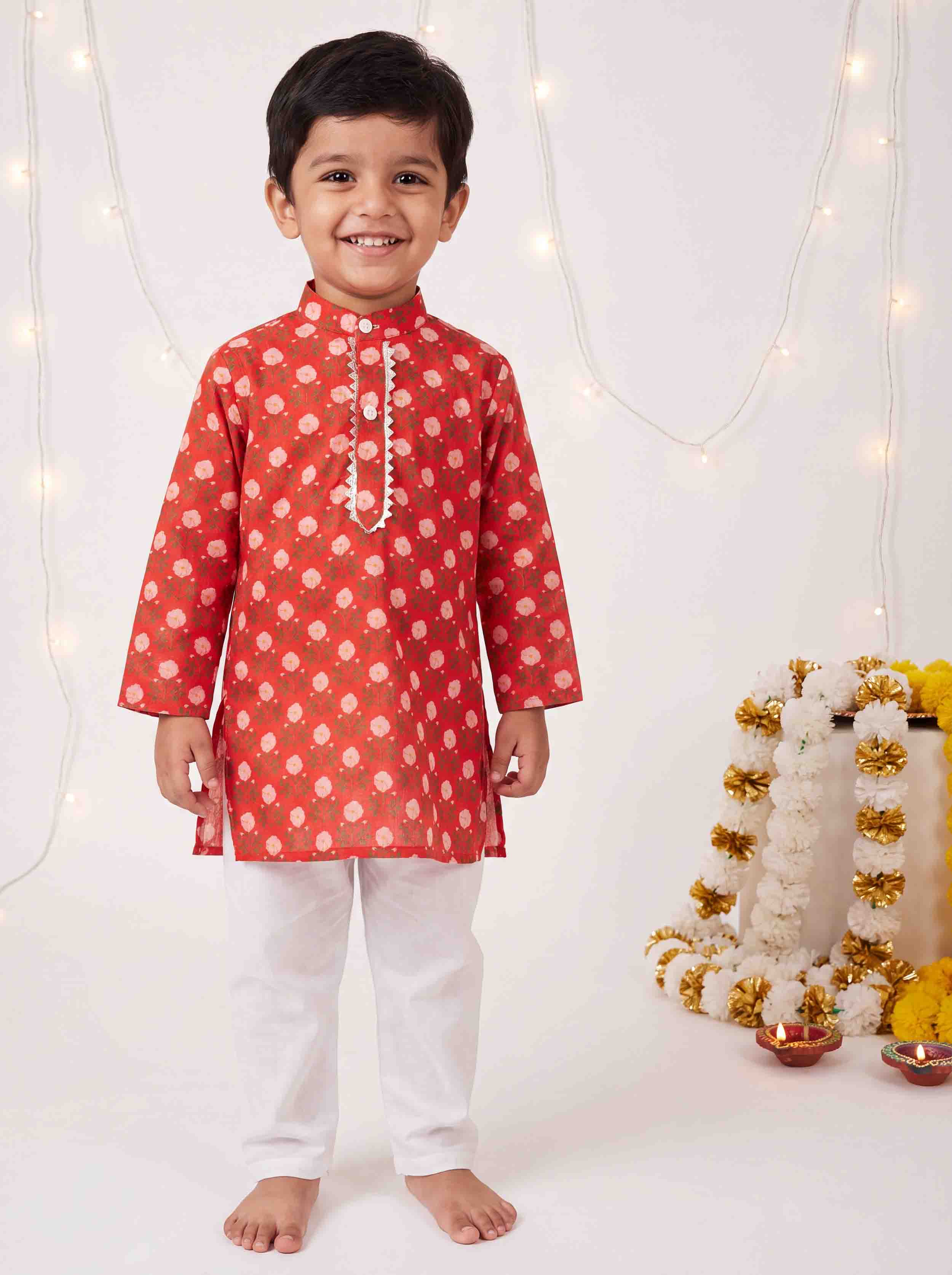 A young boy smiling and wearing a red kurta with white polka dots and white pants, standing barefoot against a decorated white background with warm fairy lights and a garland.