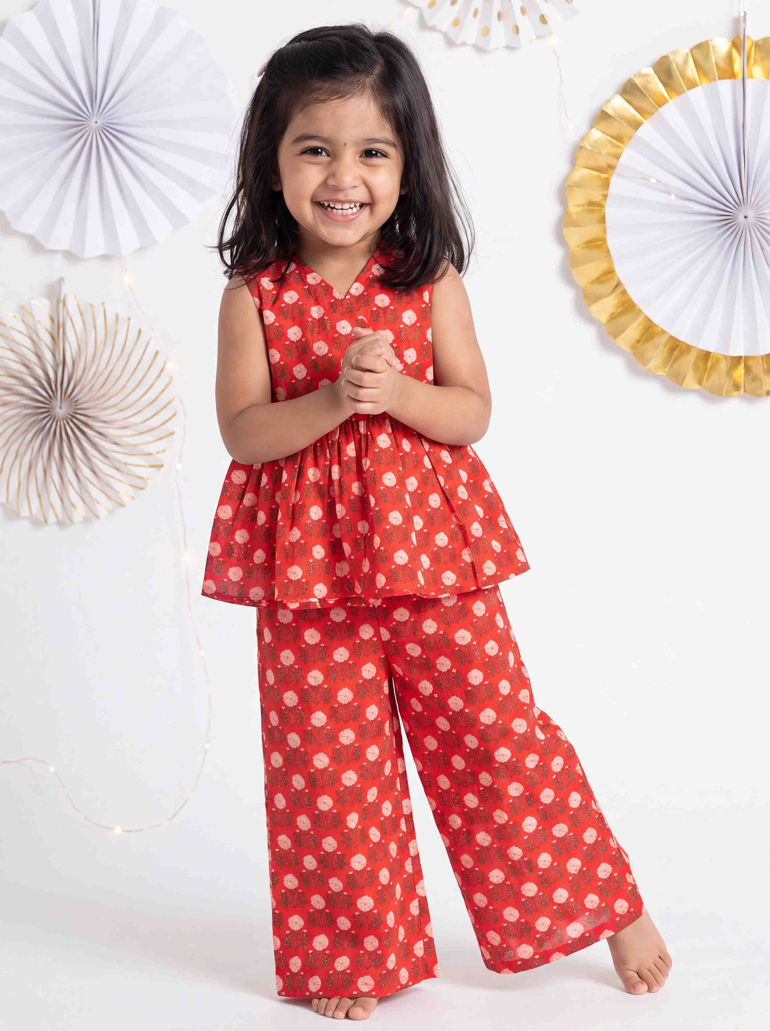 A young girl smiling and wearing a red sleeveless peplum top and matching wide-leg pants set with a white polka dot pattern, standing against a white wall decorated with paper fans.