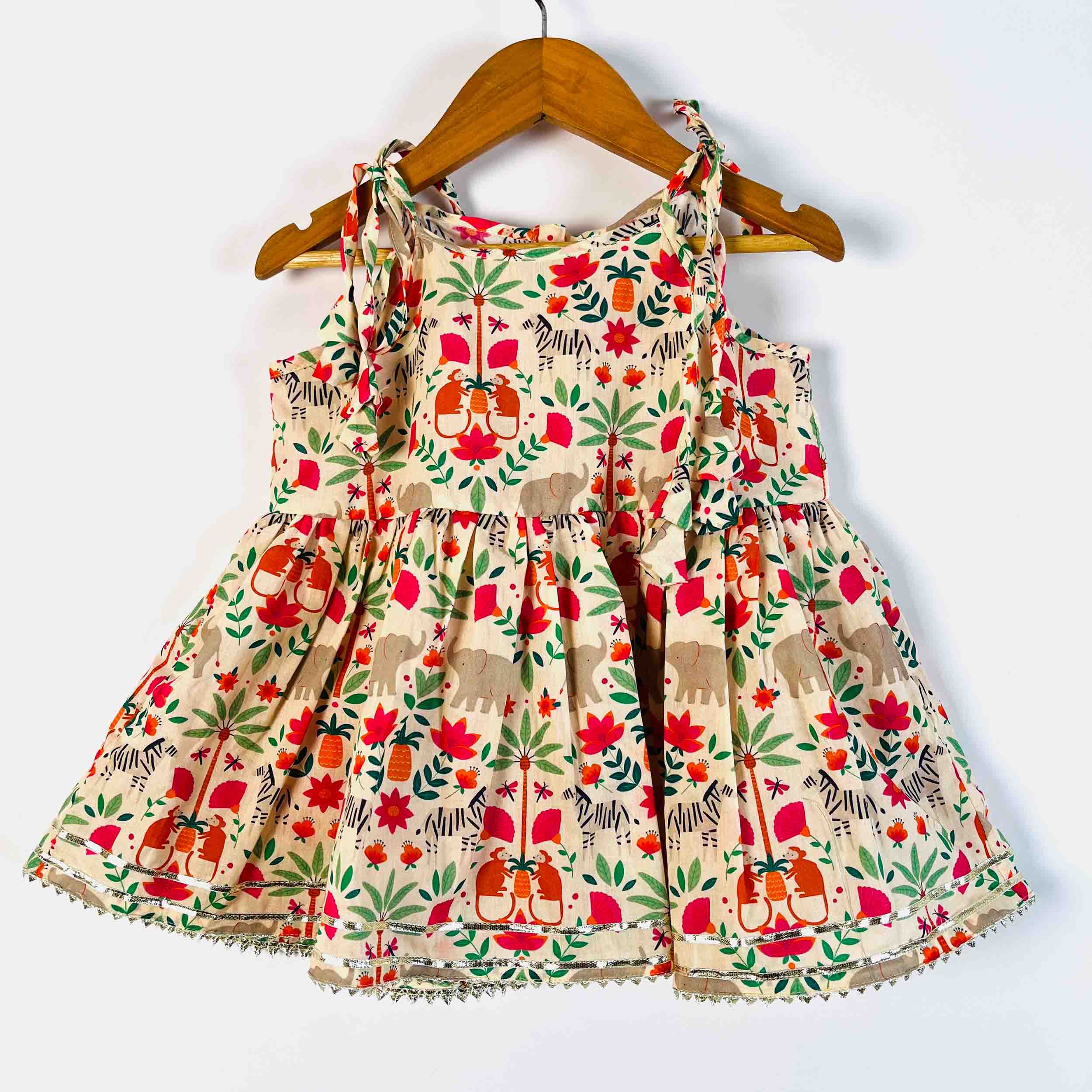 A sleeveless baby dress with a colorful safari print featuring elephants, zebras on a beige background, hanging on a wooden hanger against a plain white wall.