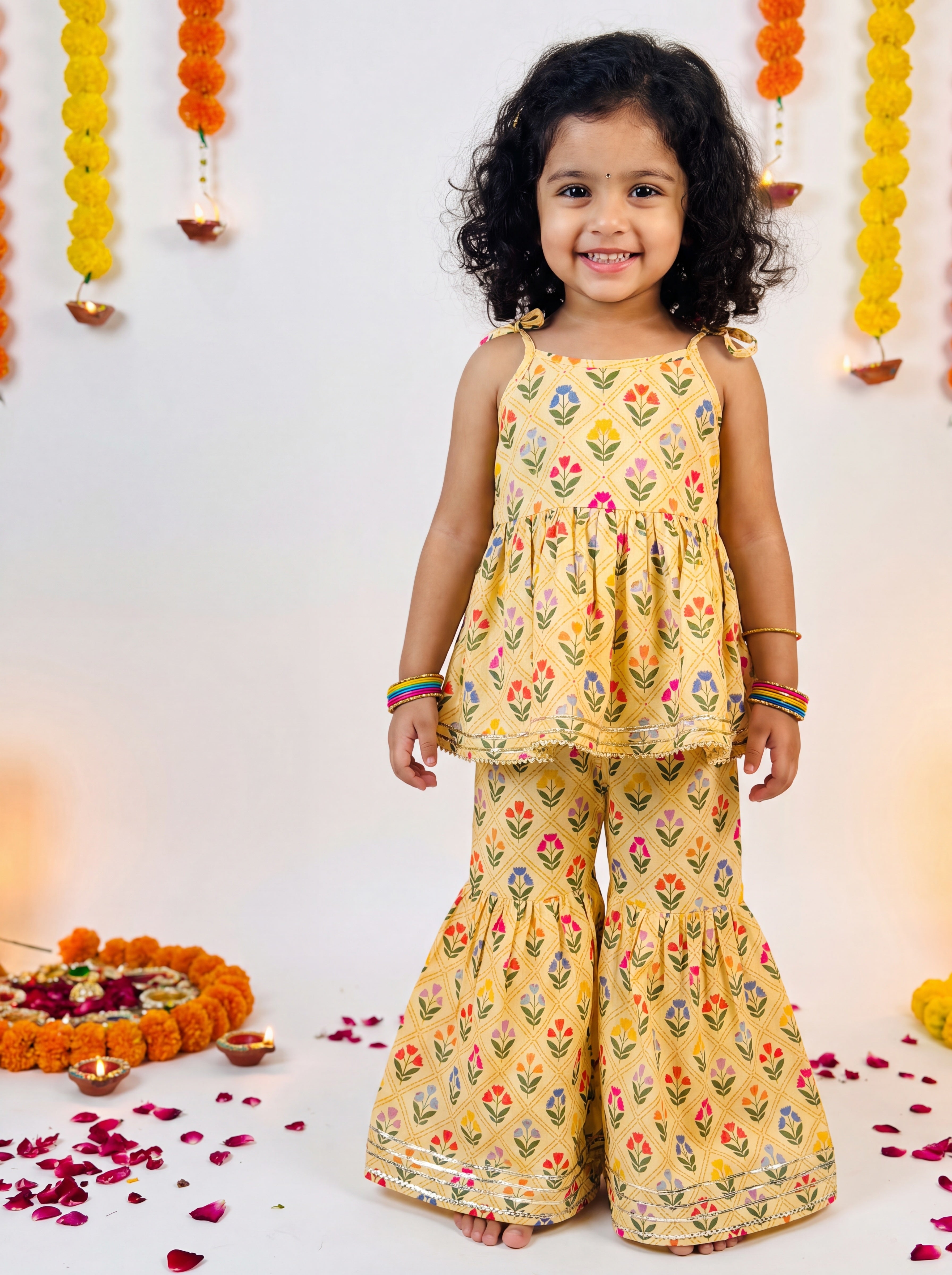 A young girl with curly black hair stands barefoot wearing a yellow sleeveless two-piece outfit with a colorful floral print. The set includes a peplum-style top and matching flared pants.