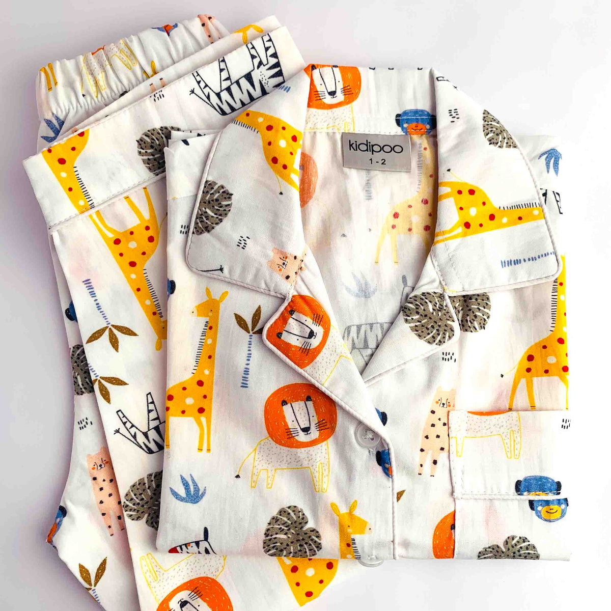 A folded children's pajama set with a playful safari-themed print featuring giraffes, lions, zebras, and trees on an off-white background. The brand label 'kidpoo' is visible on the collar.