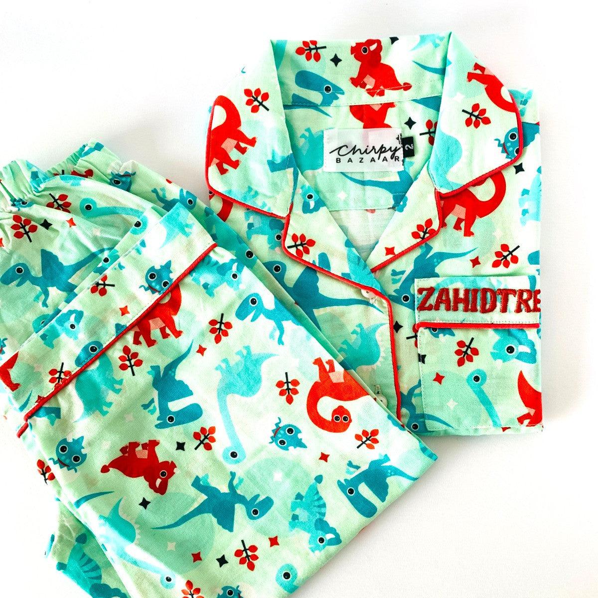 A baby night suit with dinosaur print, consisting of a long sleeve shirt and pajamas with elastic at the waist, laid flat on a white background.