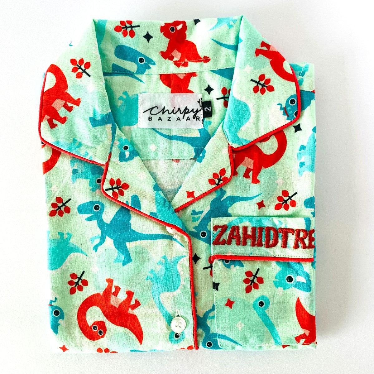 A baby night suit with dinosaur print, consisting of a long sleeve shirt and pajamas with elastic at the waist, laid flat on a white background.