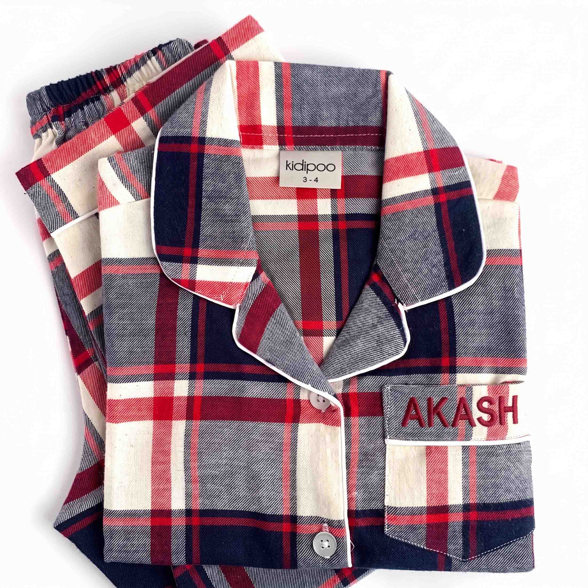 A folded plaid pajama set on a white background, featuring a long-sleeve button-up shirt with a collar and matching pants. The fabric has a plaid pattern of navy blue, red, and cream, with white piping.