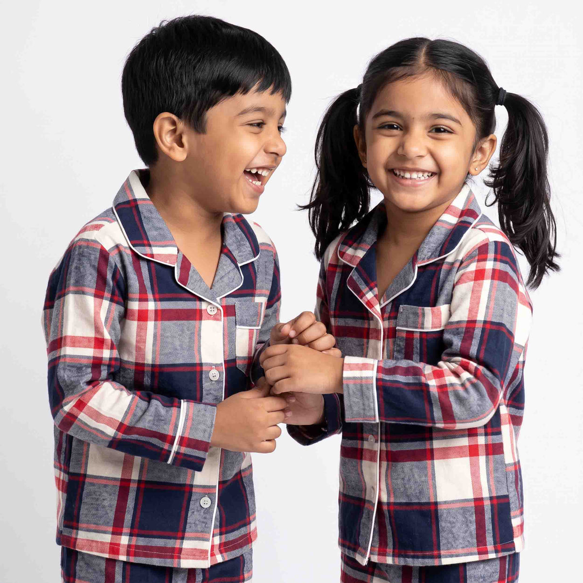 Two children wearing matching long-sleeve plaid pajamas with red, navy, and white check patterns and white piping, standing against a light gray studio background.