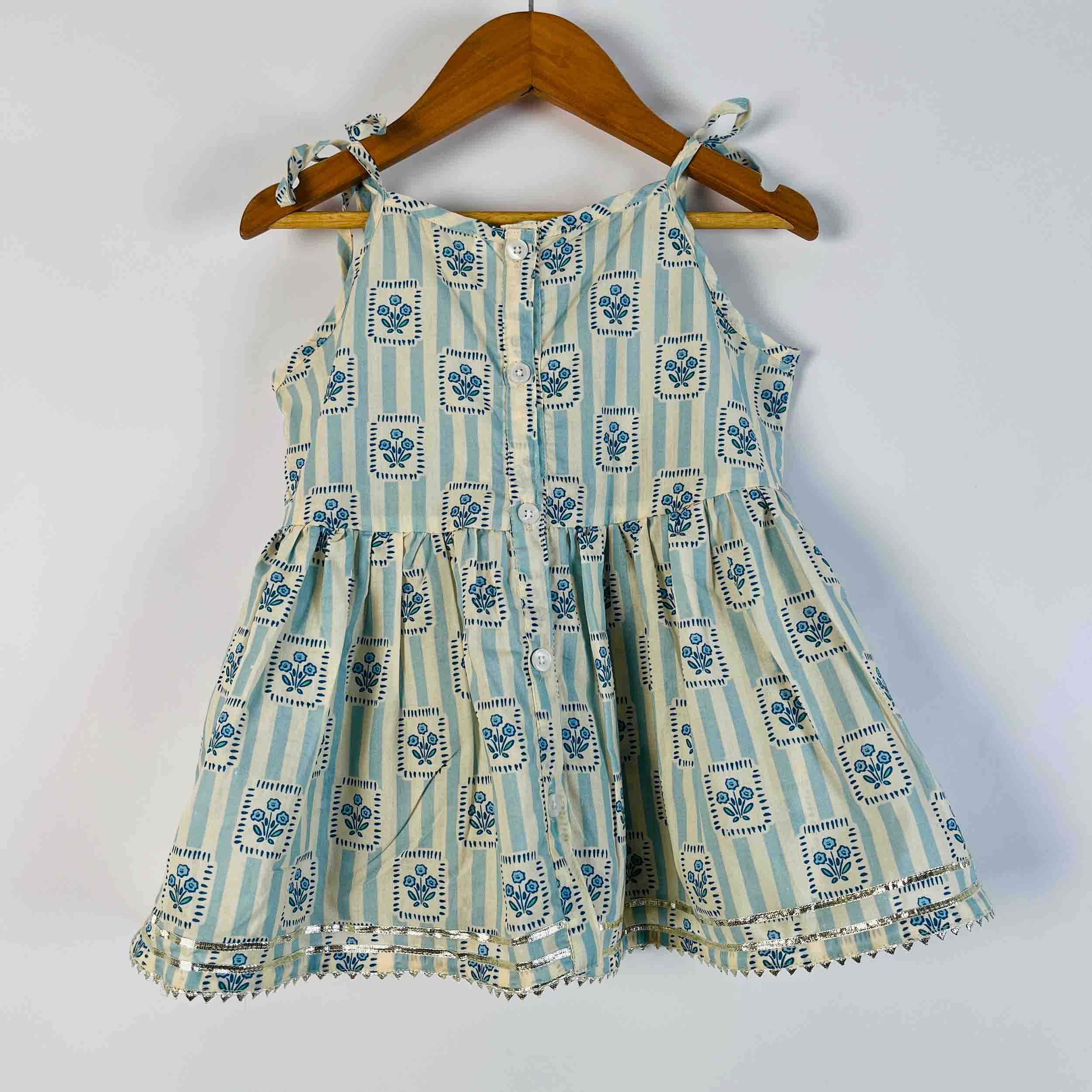 A sleeveless tiered dress for toddlers, featuring a light blue and cream vertical stripe pattern with blue floral block prints, hanging on a wooden hanger against a plain light gray wall.