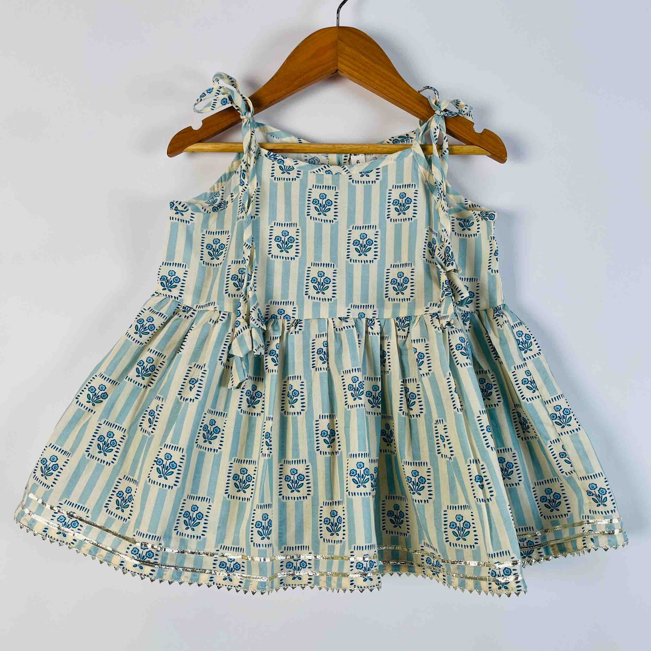 A sleeveless baby dress with a tiered skirt, featuring a light blue and white vertical stripe pattern with blue floral motifs in each stripe. The dress has tie-shoulder straps and is hanging on a wooden hanger against a plain white wall.
