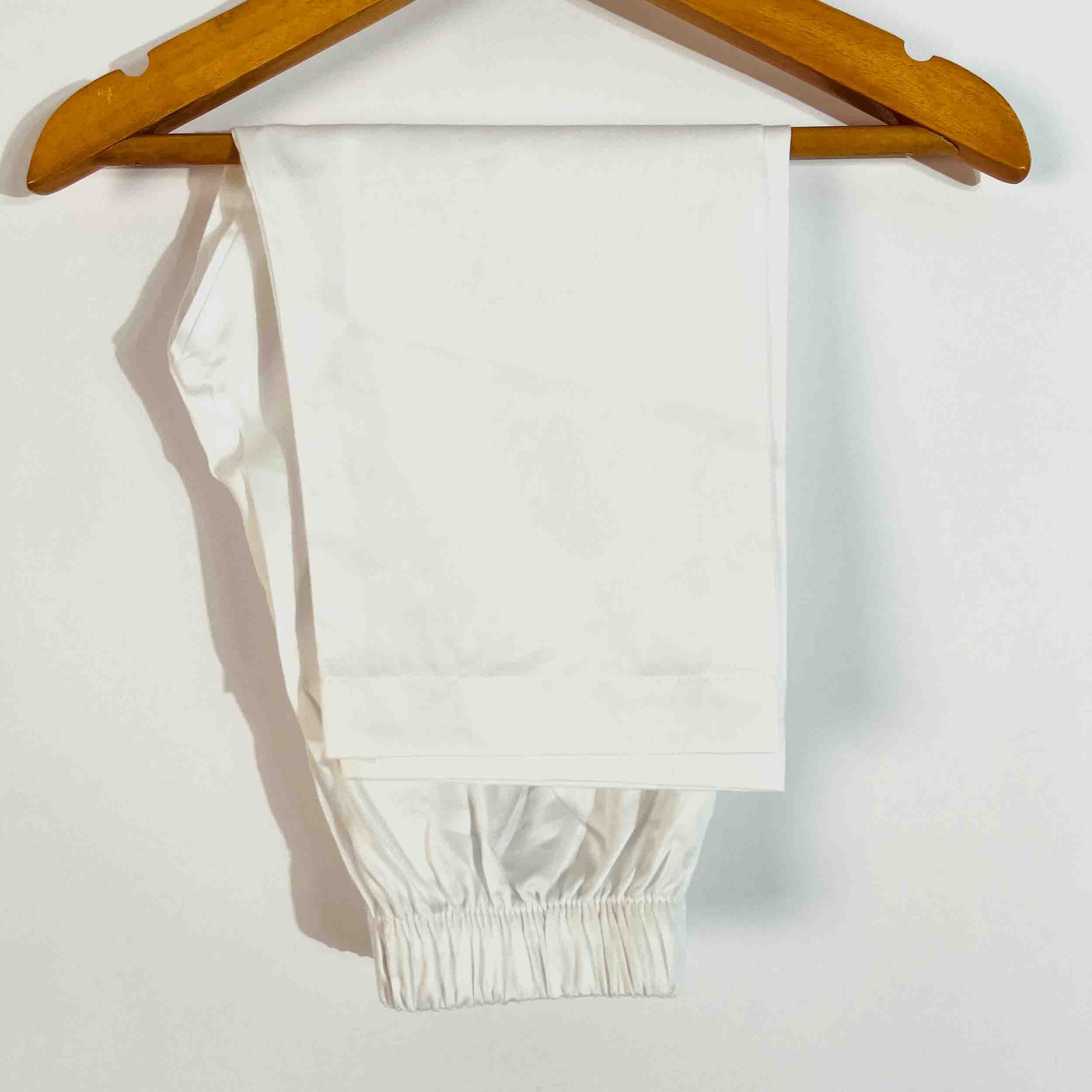 A pair of white elastic-waist pants hanging from a wooden hanger against a plain white wall.