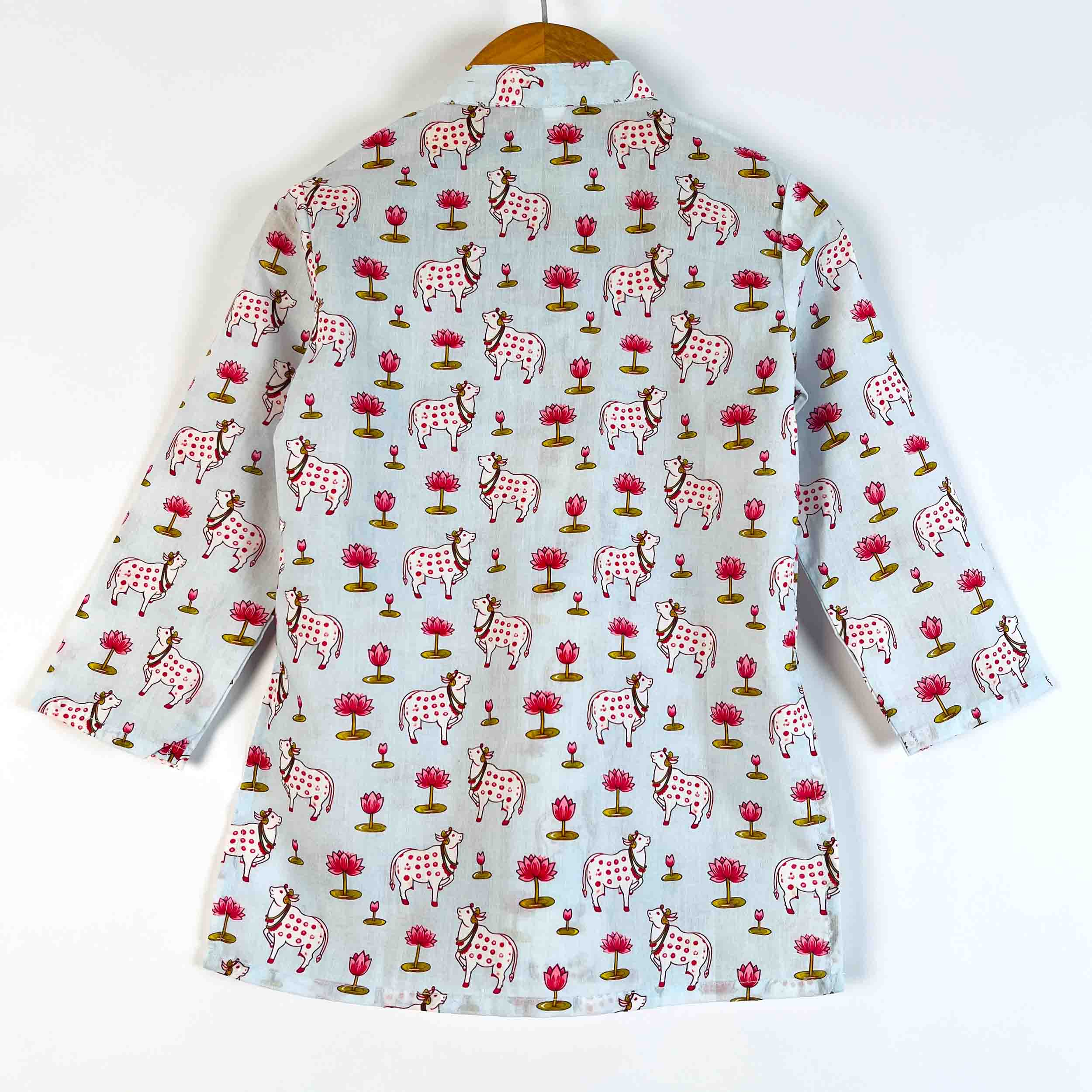 A light blue long-sleeve shirt with a repeating hand-block print of pink lotus flowers and spotted cows, hanging on a wooden hanger against a plain white background.