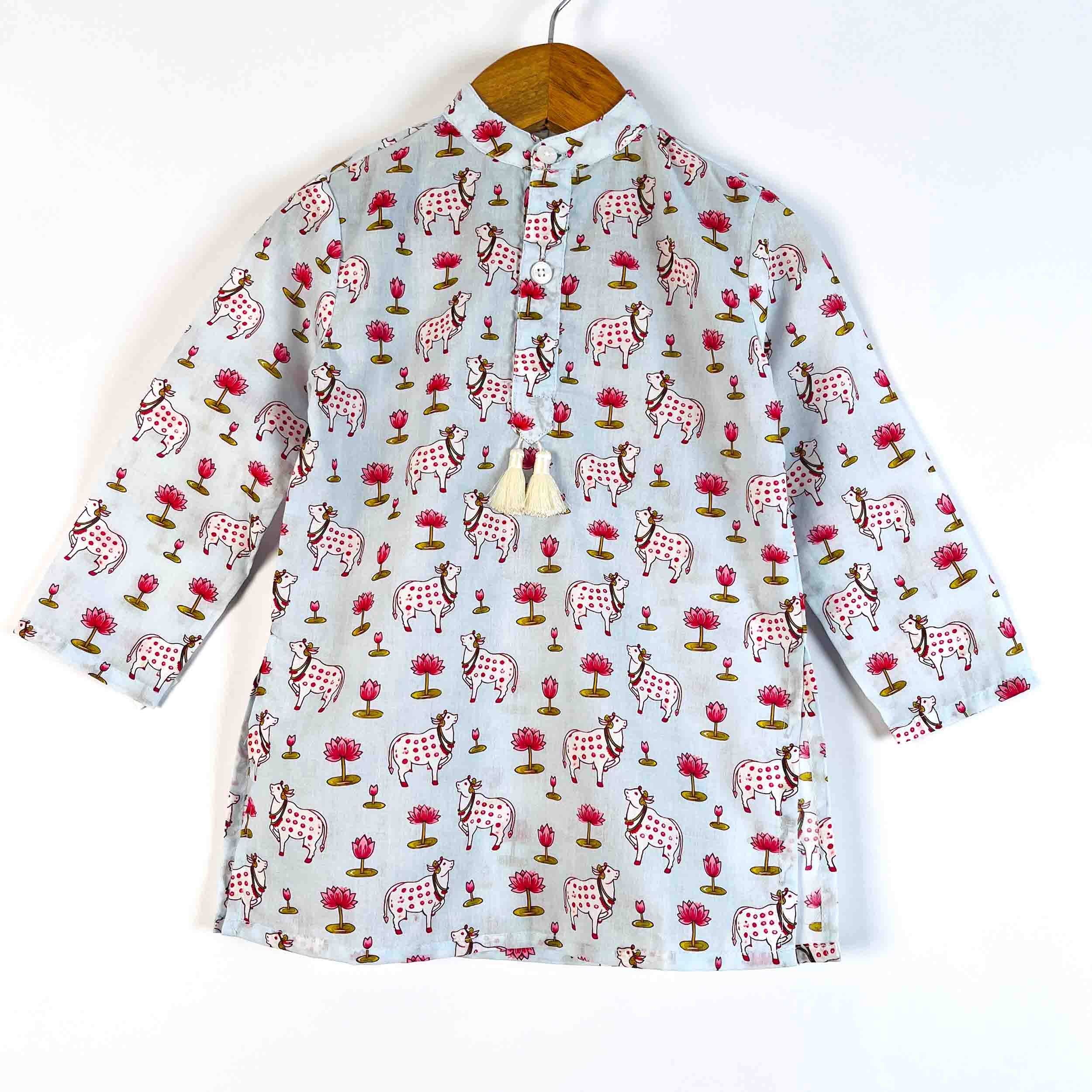 A light blue long-sleeve shirt with a colorful hand-block print of cows, trees, and flowers, hanging on a wooden hanger against a white background.