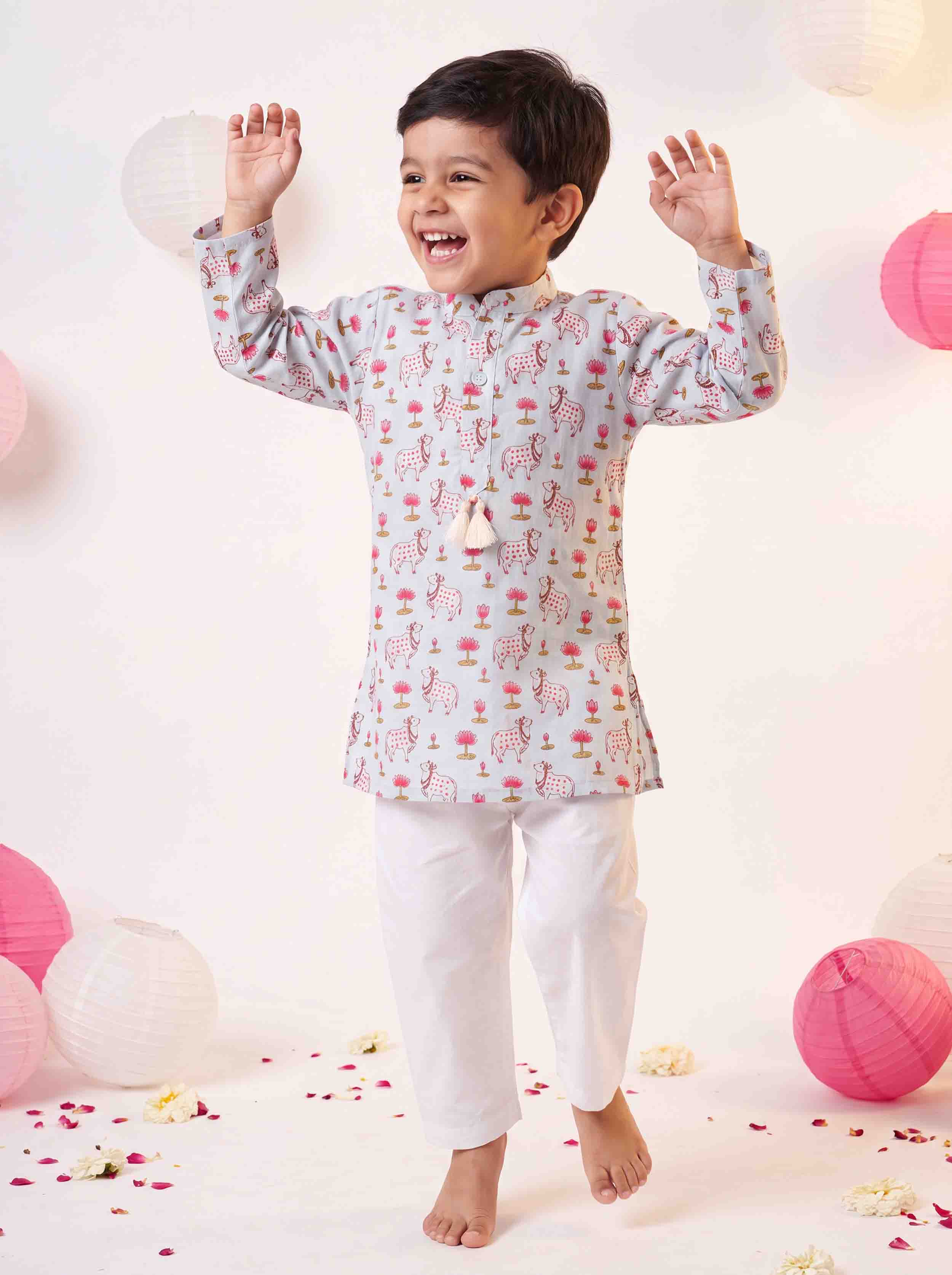 A young boy wearing a light gray and pink printed kurta with white pants, standing barefoot in a studio decorated with paper lanterns and flower petals.