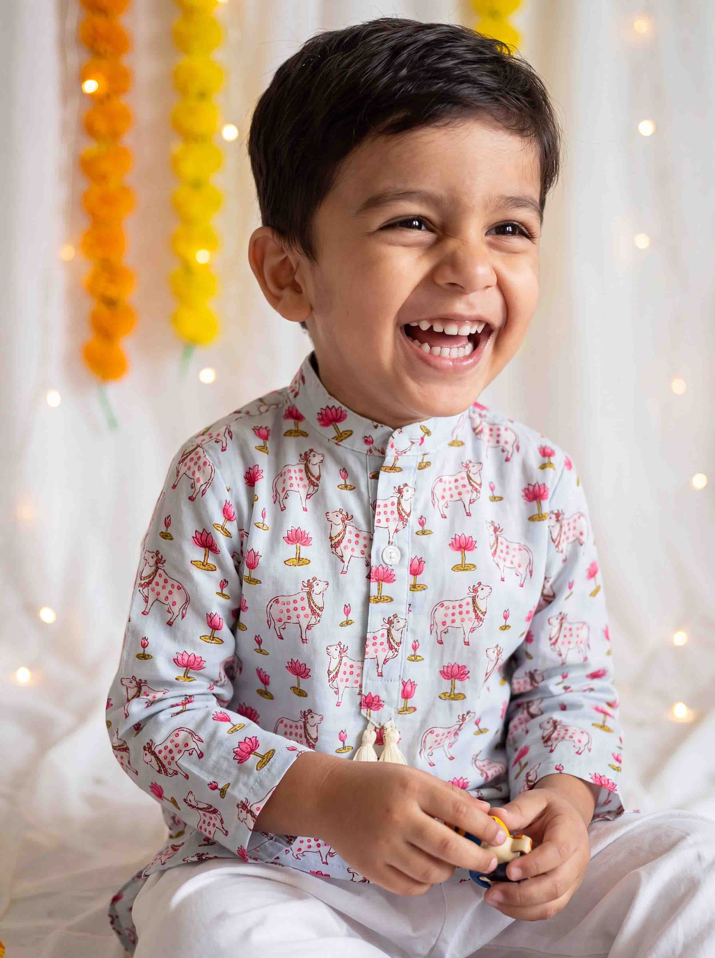 A young boy laughing happily while wearing a light gray long-sleeve kurta with a pink and green elephant print, paired with white trousers, set against a decorated white curtain background with yellow marigold garlands.