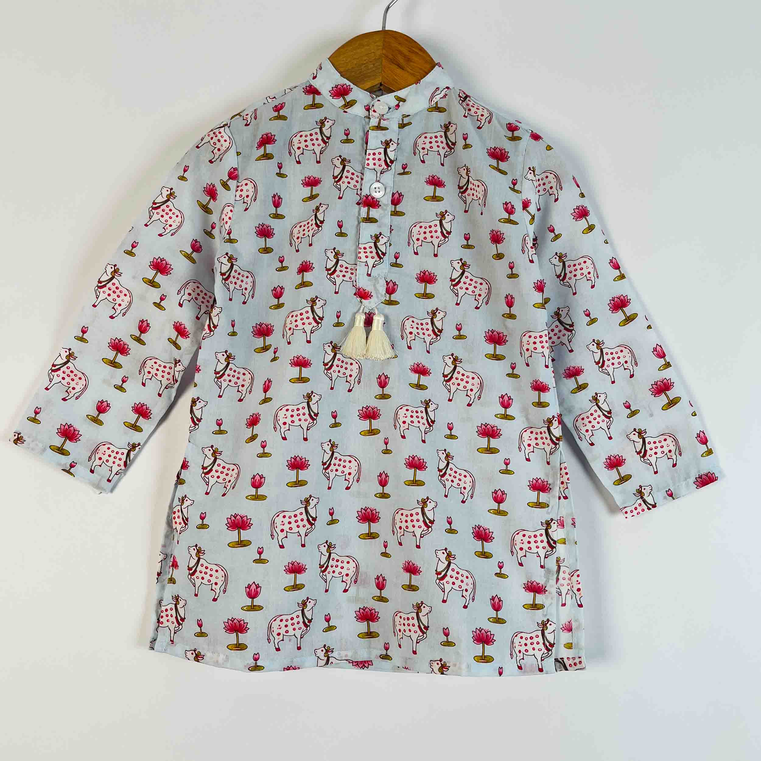 A light gray long-sleeve shirt with a colorful print of pink and white spotted cows, red flowers, and green trees, hanging on a wooden hanger against a plain white wall.
