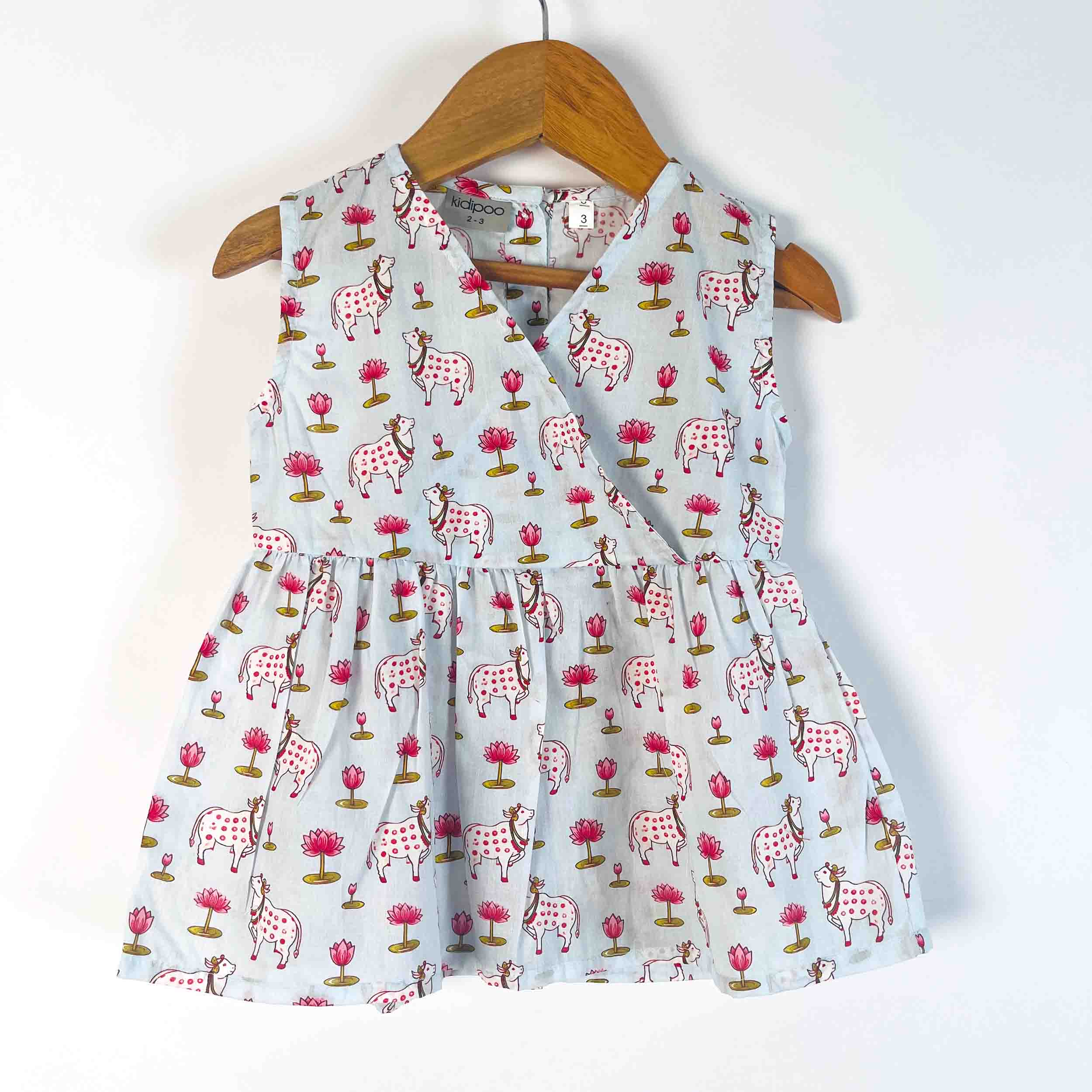 A sleeveless baby dress with a light blue-gray background and an all-over print of pink flowers and spotted horses, hanging on a wooden hanger against a plain white wall.