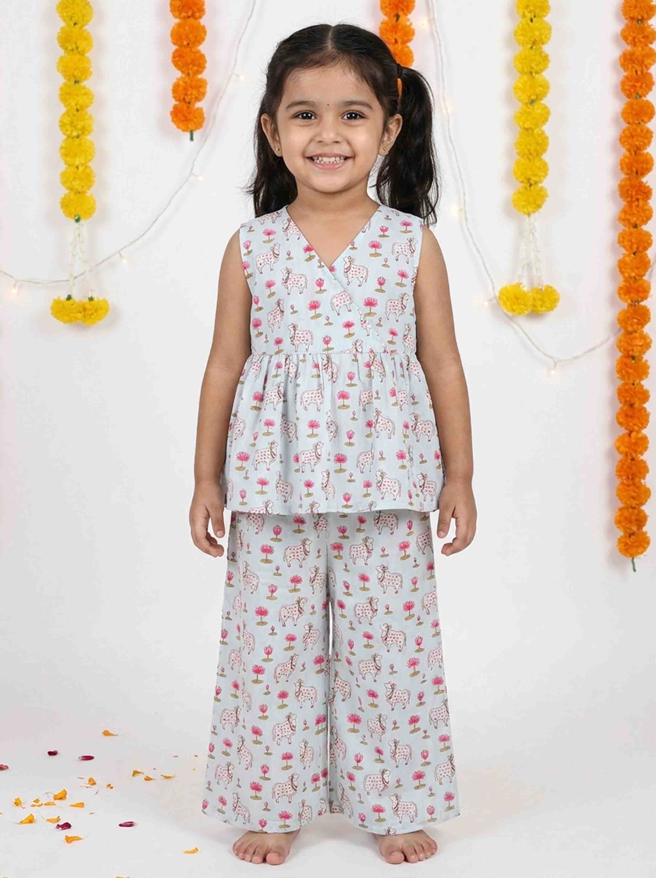 A young girl stands barefoot against a white backdrop decorated with orange and yellow marigold garlands. She is wearing a sleeveless V-neck tunic top and matching wide-leg pants in a light blue-gray fabric with a pink and red floral print.