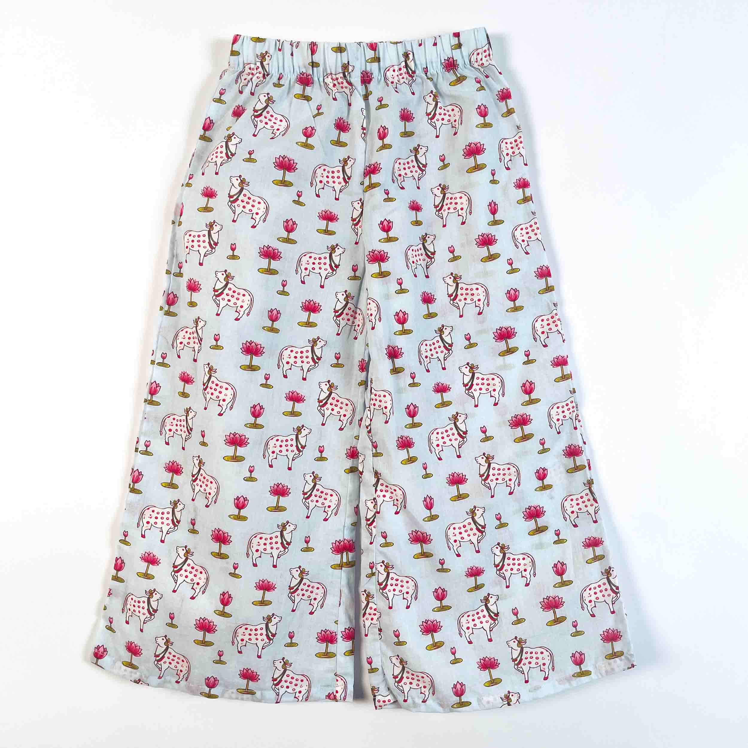A pair of wide-leg shorts with an all-over print of pink and white spotted cows, pink flowers, and green trees on a light gray background.