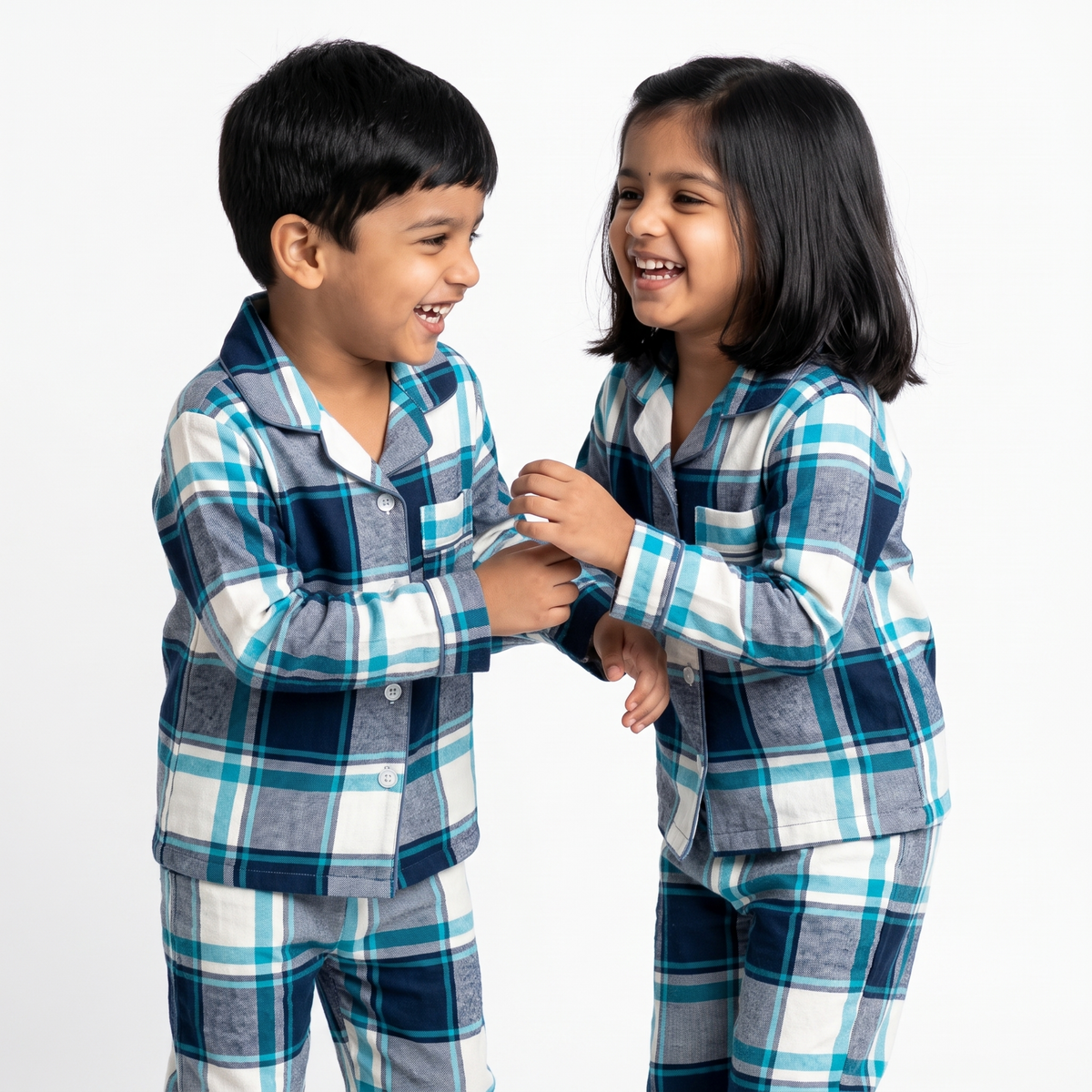Two children wearing matching long-sleeve plaid pajamas in navy, teal, gray, and white, standing and smiling at each other against a plain white studio background.