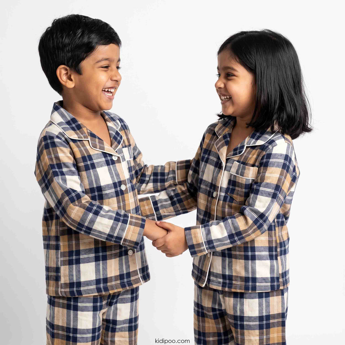Two children smiling and holding hands while wearing matching plaid pajamas in beige, navy, and white, photographed against a clean light gray studio background.