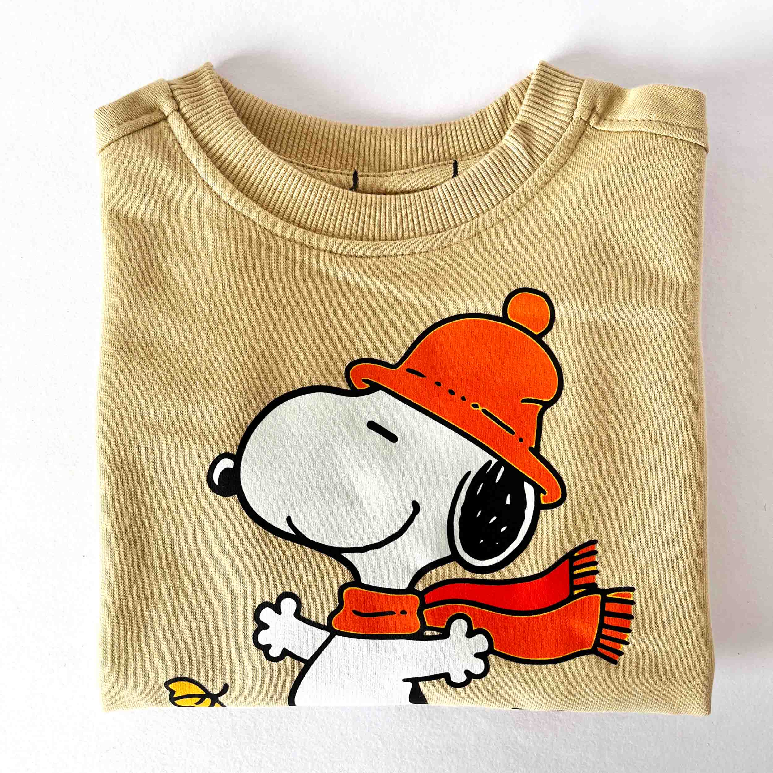 Folded beige sweatshirt with a cartoon character wearing an orange hat and red scarf on a white background