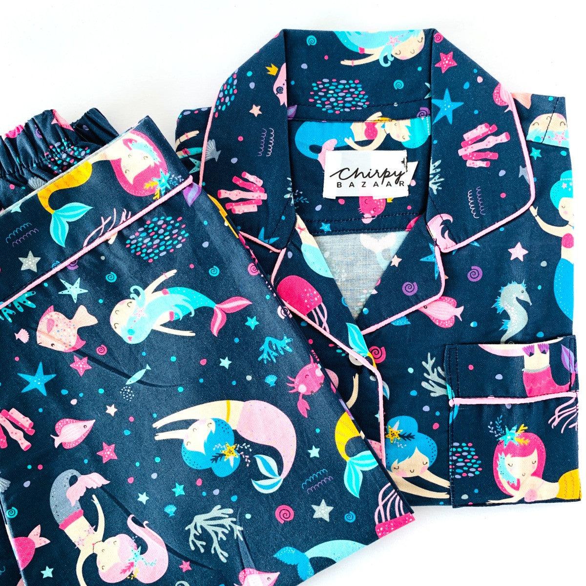 A set of children's sleepwear featuring a long sleeve shirt and pajamas with elastic at the waist, decorated with colorful dancing mermaids and stars on a dark blue background.