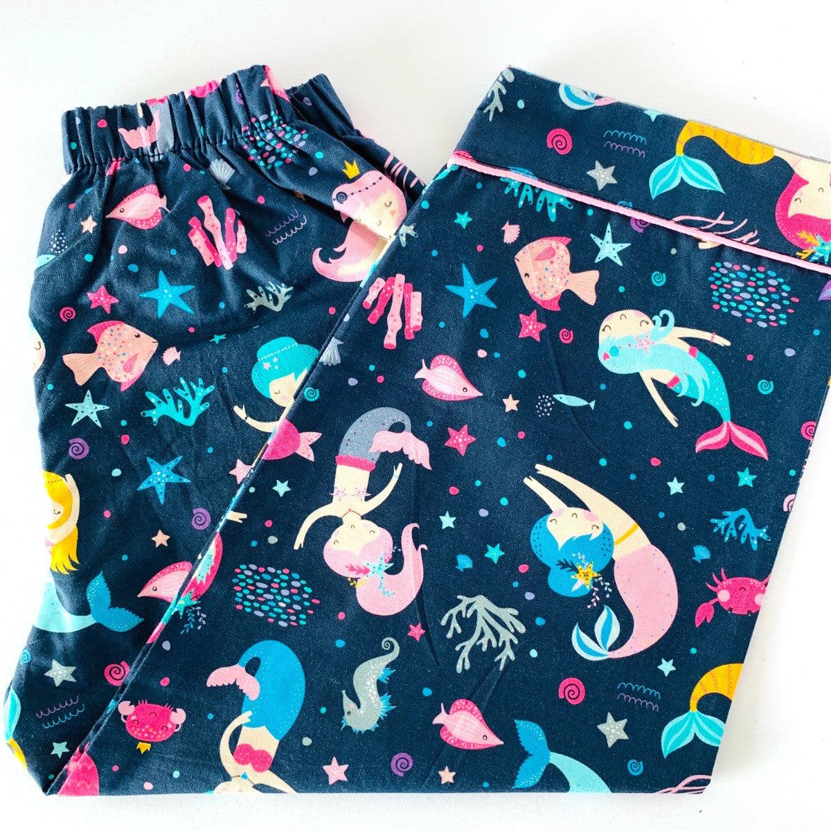 A set of children's sleepwear featuring a long sleeve shirt and pajamas with elastic at the waist, decorated with colorful dancing mermaids and stars 