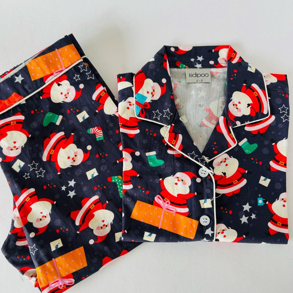 A night suit with a long sleeve, front button shirt and coordinated pajamas with elastic at waist, featuring a Christmas themed pattern with Santa Claus, Christmas trees, and holiday symbols.