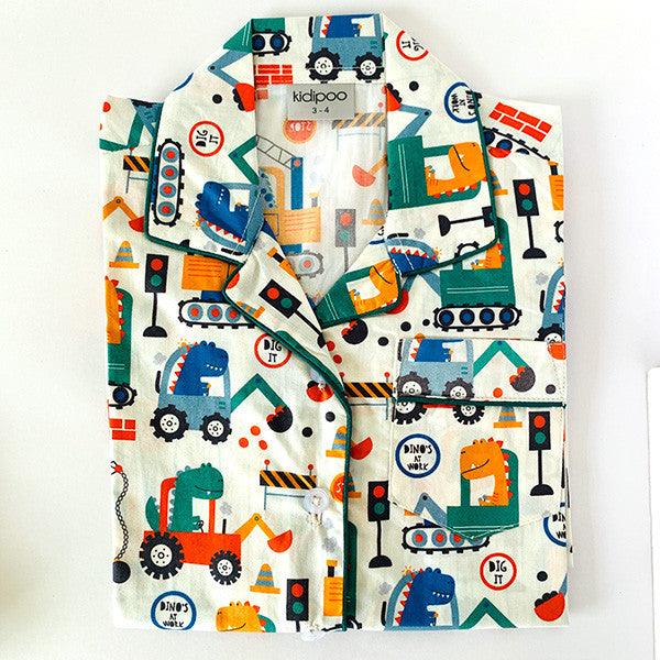 A set of children's pajamas with a dinosaur theme, including a long sleeve, front button shirt and coordinated pajamas with elastic at the waist, laid out on a white surface.