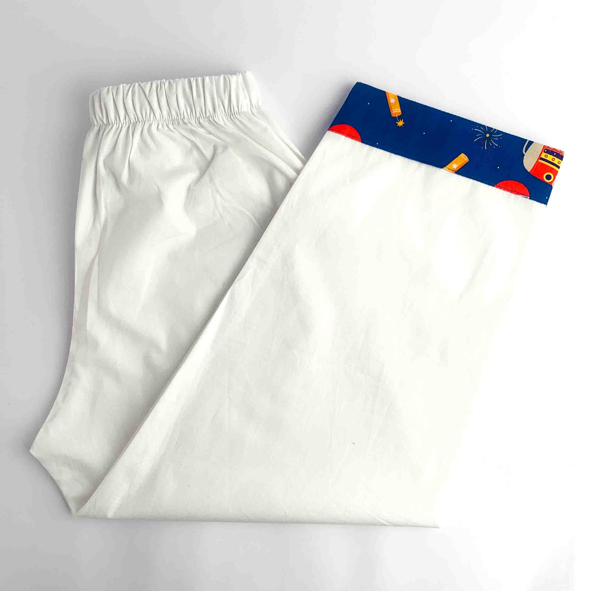 A pair of white cotton pajama pants with an elastic waistband, folded on a white background. One leg is unfolded to show a decorative waistband featuring a navy blue space-themed print with red planets, yellow rockets, and white stars.
