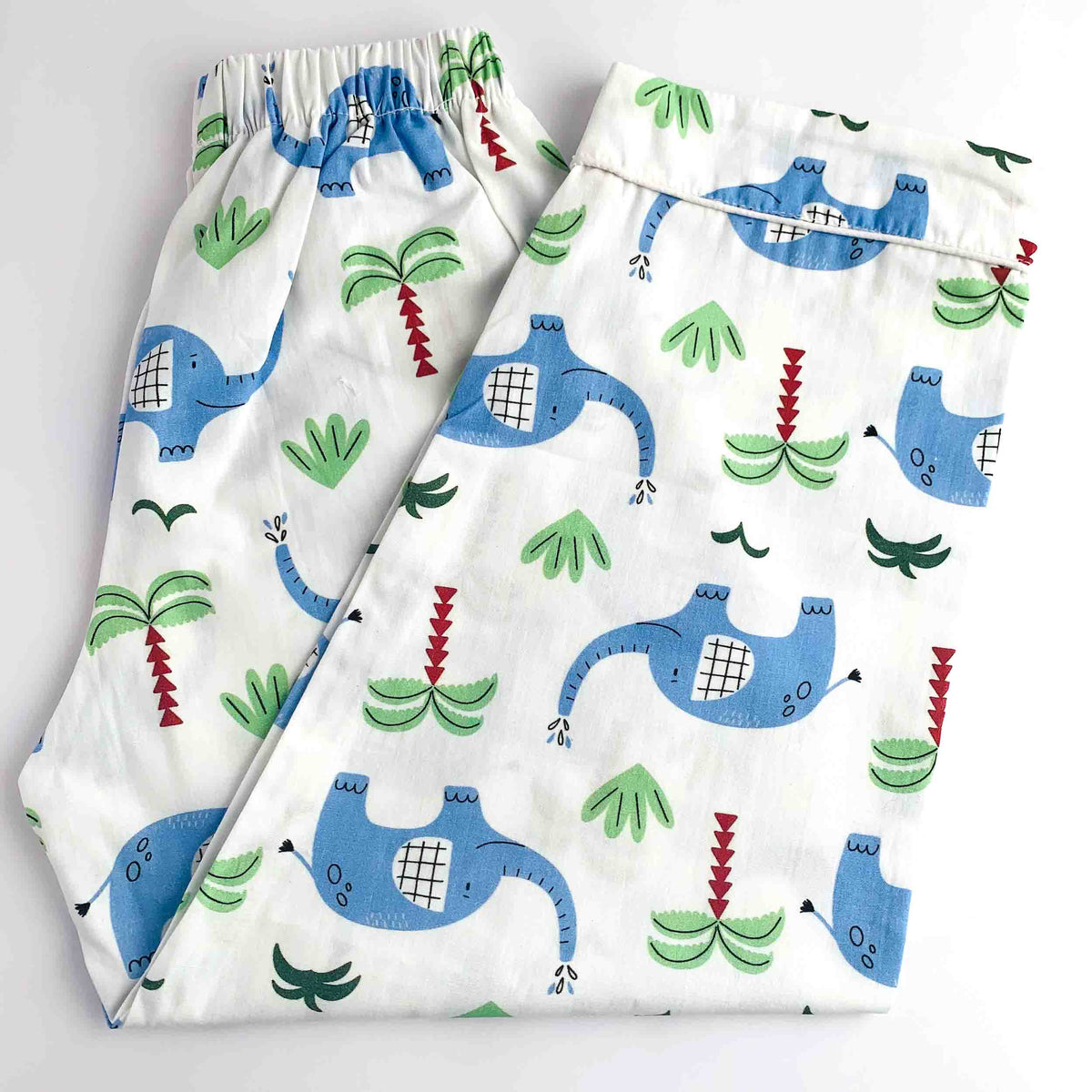 A pair of white children's pajama pants with an elastic waistband, featuring a playful cartoon print of blue dinosaurs, green palm trees, and red plants.