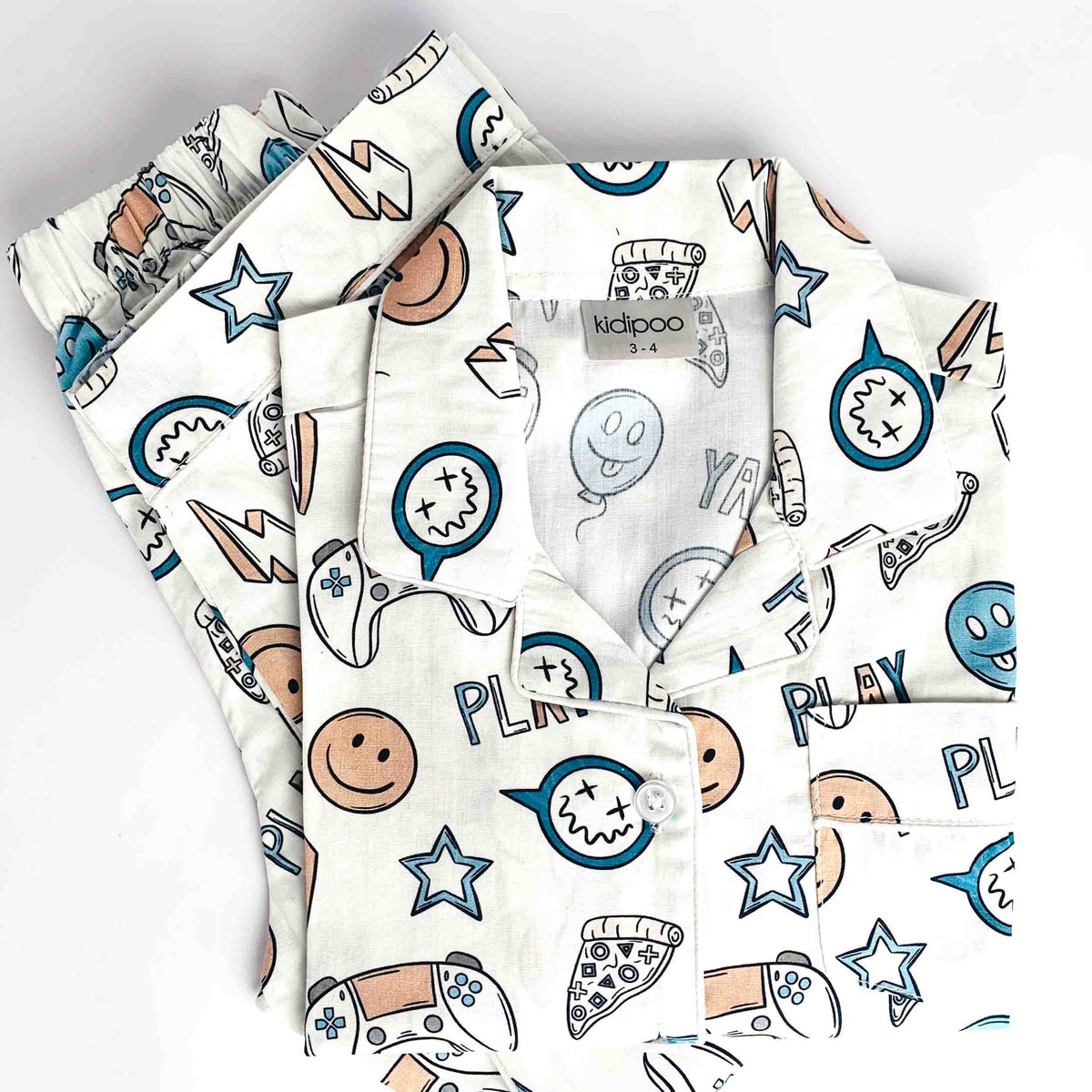 A folded white pajama shirt with a colorful cartoon print featuring stars, smiley faces, pizza slices, game controllers, and text like 'PLAY' and 'RULY', shown on a plain white background.