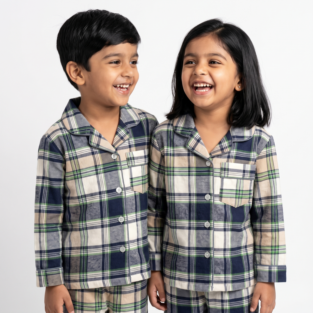 Two children wearing matching long-sleeve plaid pajamas with a navy, green, and beige check pattern, standing against a plain light gray background.