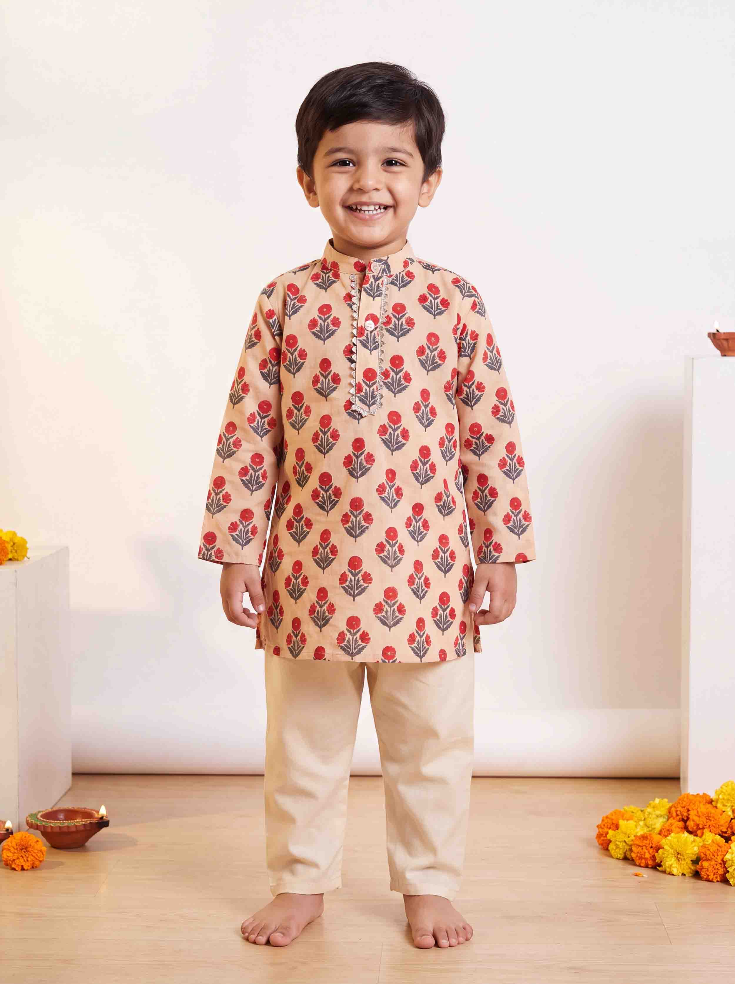 A young boy smiling and standing barefoot indoors, wearing a beige kurta with a red and gray floral block print and beige trousers.