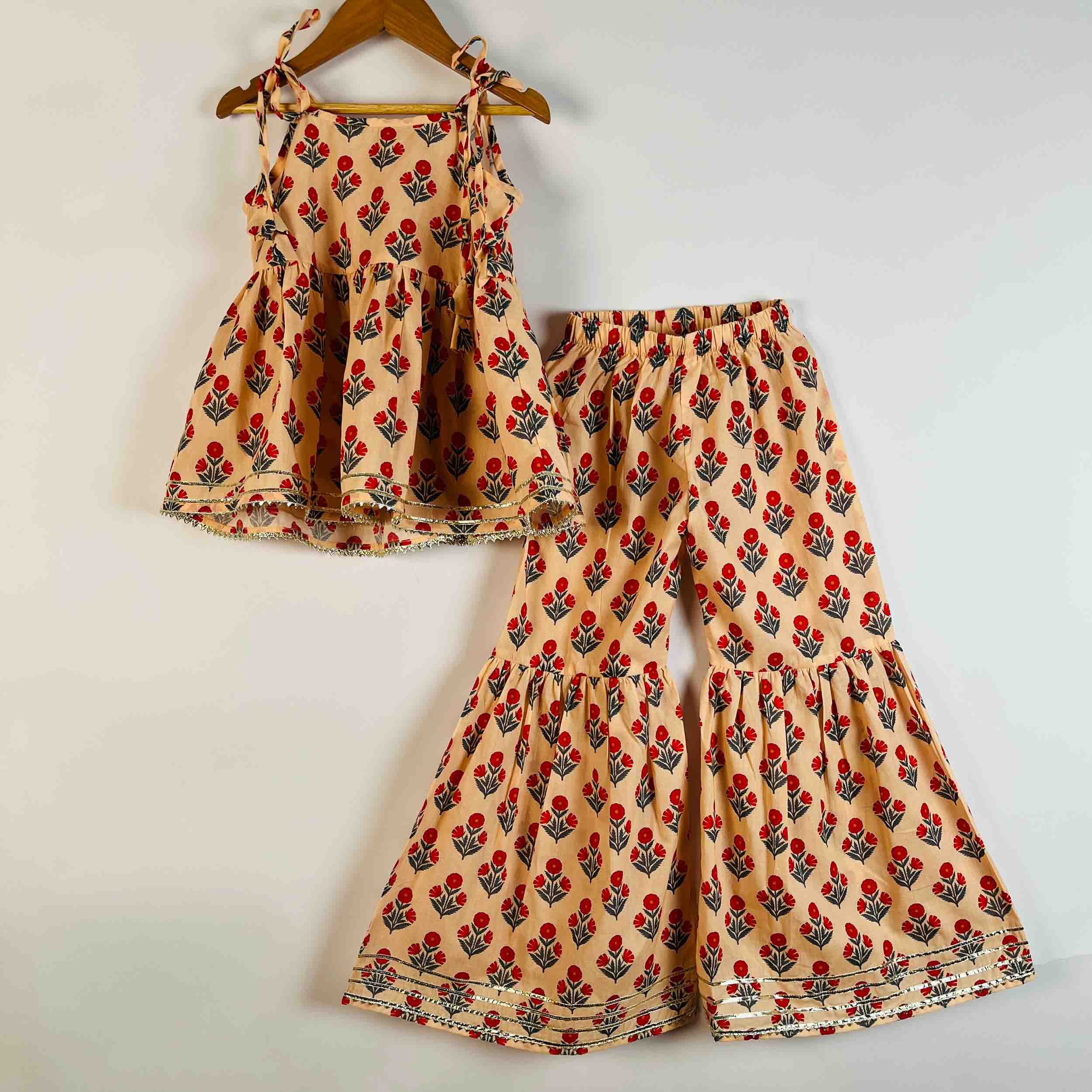 A two-piece children's outfit set hanging on a wooden hanger against a plain light background. The set includes a sleeveless peplum-style top with shoulder ties and matching wide-leg flared pants, both in a warm beige fabric with an allover red and green floral block print.