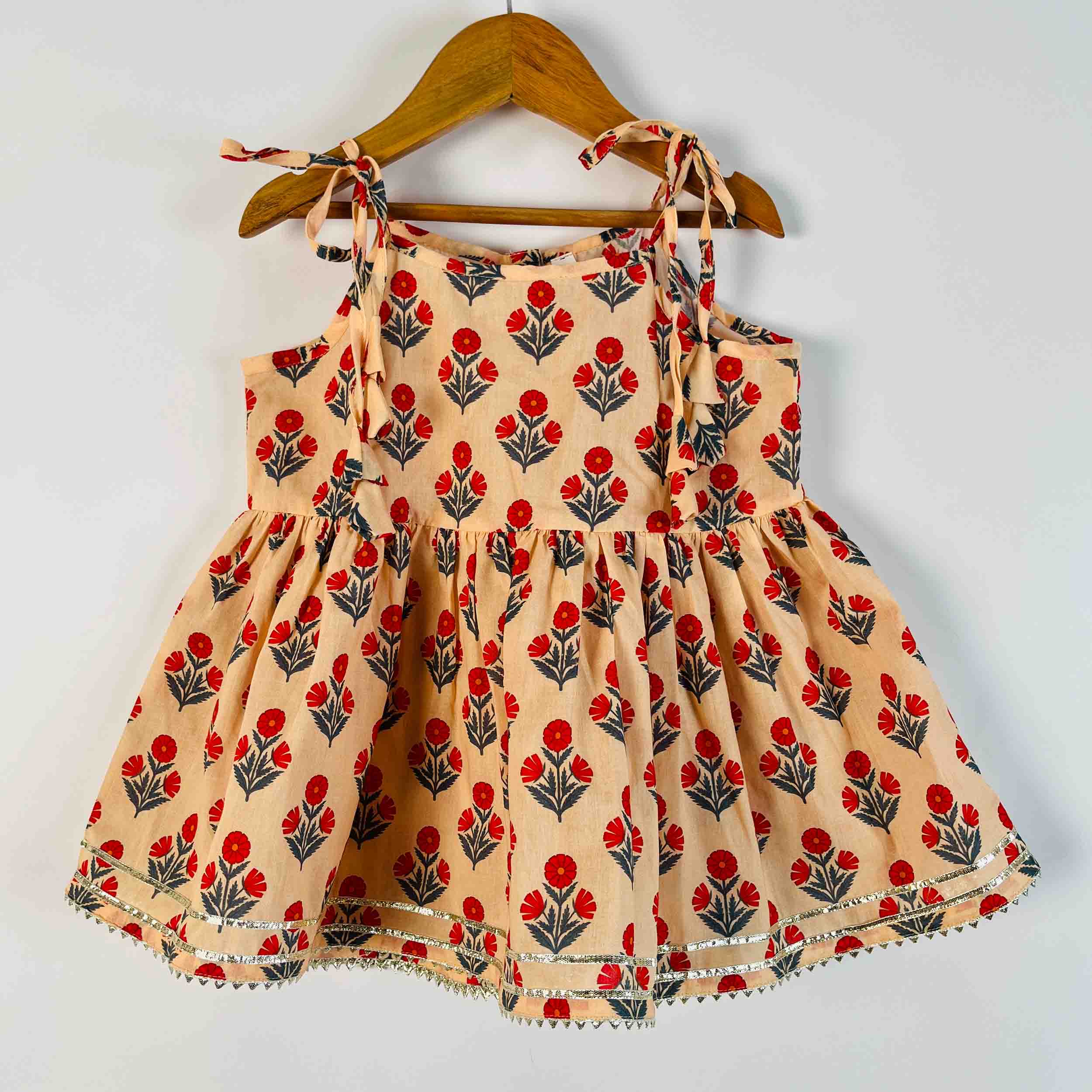 A sleeveless baby girl's dress with a red and green floral block print on a beige background, hanging on a wooden hanger against a plain white wall.