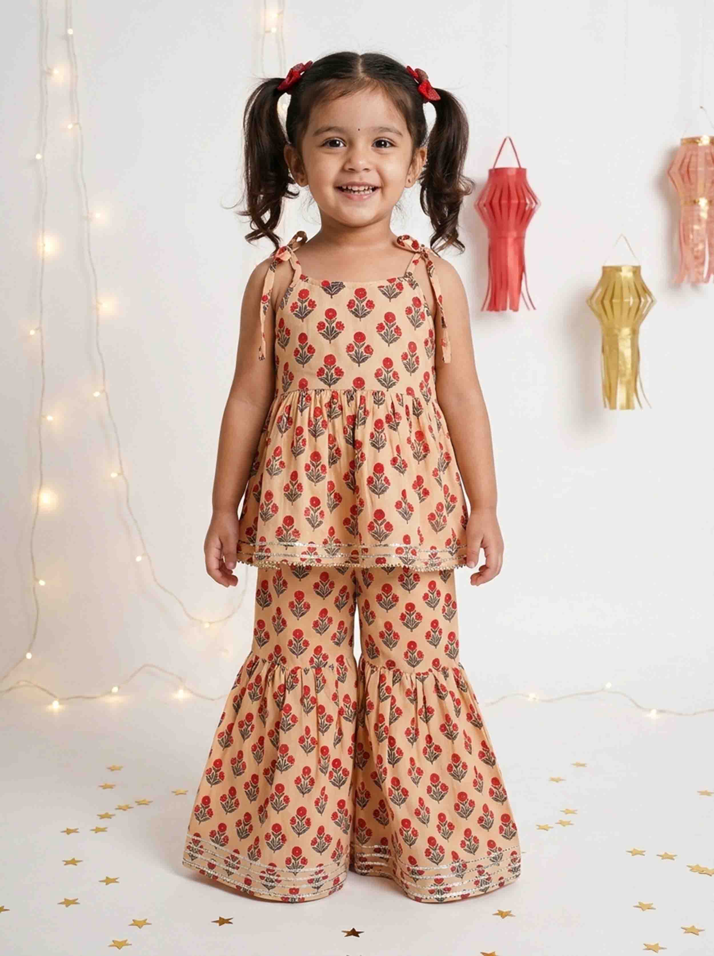 A young girl with dark hair in pigtails stands smiling, wearing a two-piece peach-colored outfit with a red and green floral pattern. The set includes a sleeveless top and matching flared pants.