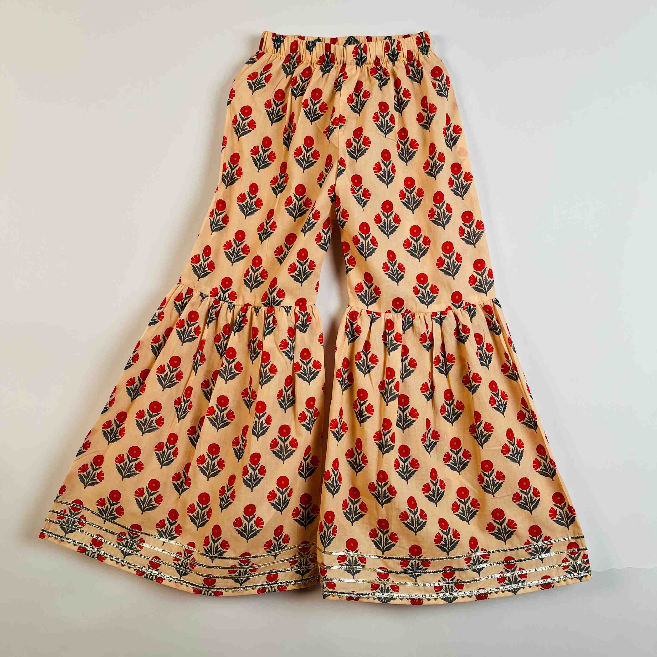A pair of wide-leg, tiered pants in beige fabric with an all-over red and green floral block print, laid flat against a light gray background.