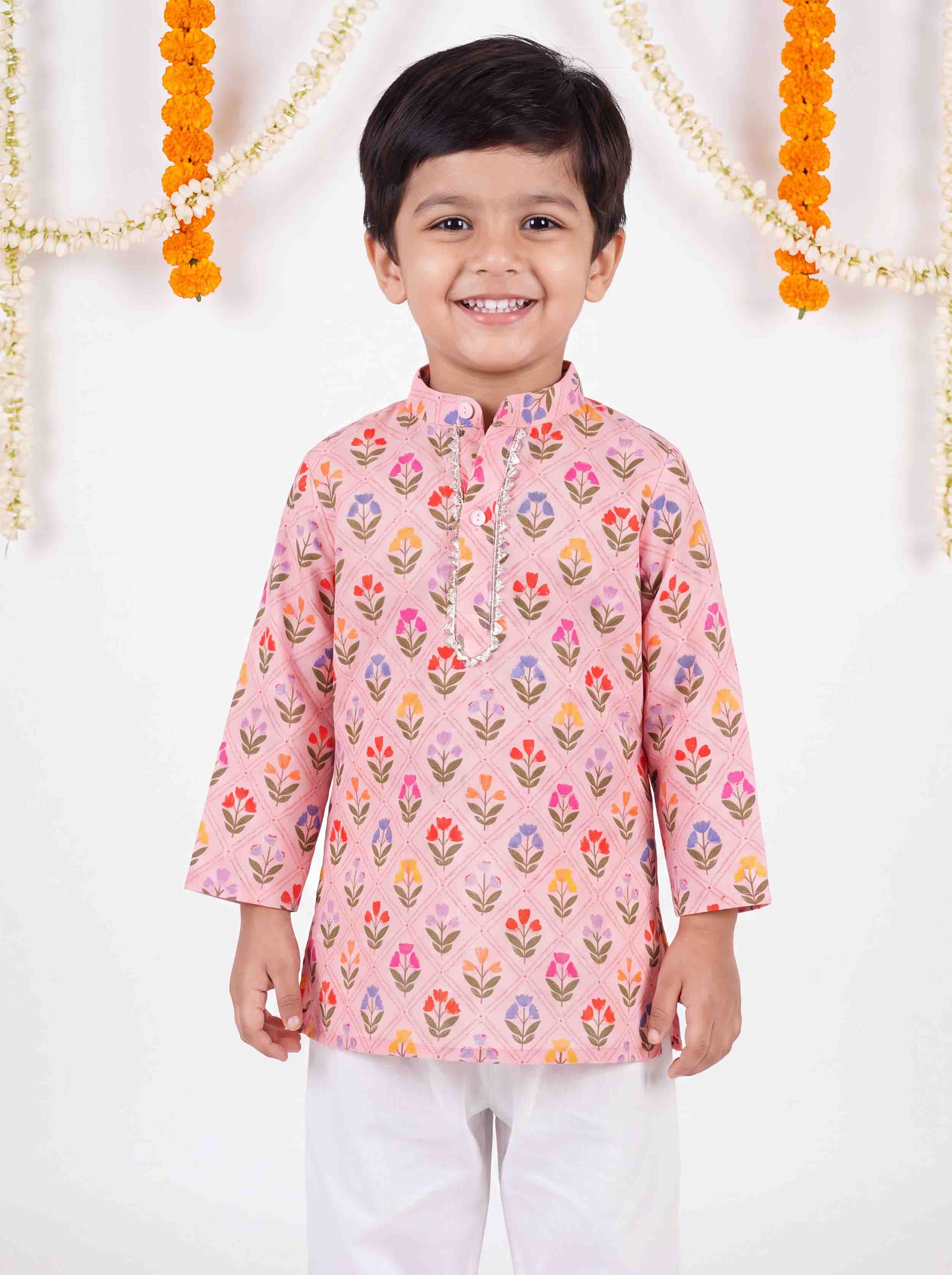 A young boy smiling and wearing a pink long-sleeve kurta with a colorful floral print, paired with white trousers, standing against a decorated white background with marigold garlands.